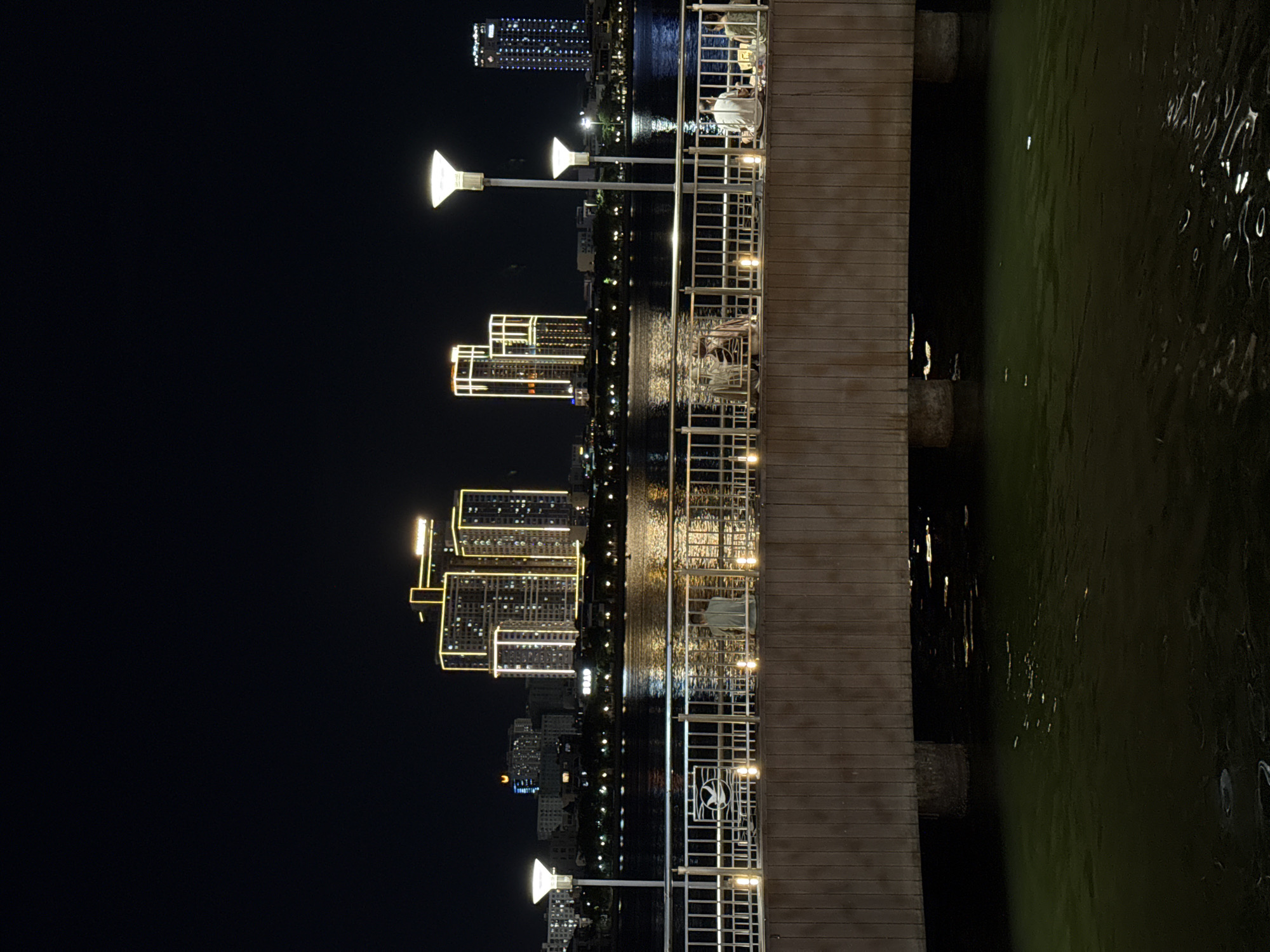 This image depicts a nighttime scene along a waterfront in Da Nang, Vietnam. The perspective is rotated 90 degrees counter-clockwise. On the right side of the frame, a wooden boardwalk with metal railings runs parallel to the water. Lights are visible along the railing, illuminating the pathway. One person is seated at a table on the boardwalk, looking towards the water.
To the left of the boardwalk, a body of water, likely a river or bay, is visible. The water reflects the lights from the buildings and the boardwalk, creating shimmering patterns.
Across the water, a cityscape of modern buildings is illuminated. Several tall buildings are prominent, with their windows and exterior lighting creating a dazzling display against the dark night sky. The buildings are brightly lit with yellow and white lights, outlining their architectural features.
Further in the background, more city lights are visible, suggesting a bustling urban environment. The overall atmosphere is one of a vibrant city at night, with the waterfront area offering a scenic viewpoint. The time of day is clearly night, and the water appears calm. The scene suggests a relaxed evening ambiance. There is no visible text in the image.