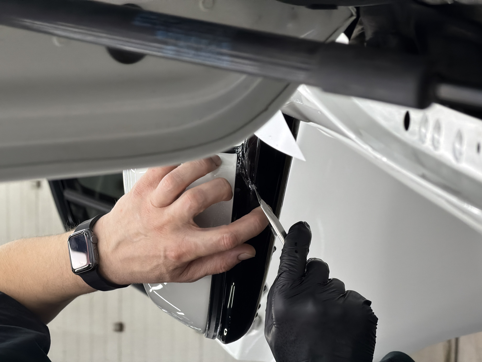 A close-up shot shows a person installing car wrap on a white vehicle. The person wears a black smart watch on their left wrist and a black glove on their right hand, which is holding a metal tool. Their bare left hand is pressing the white wrap around a black trim on the car. The background is a blurred mix of white surfaces and black components of the vehicle. No information indicates the location is Krasnoyarsk, Russia.