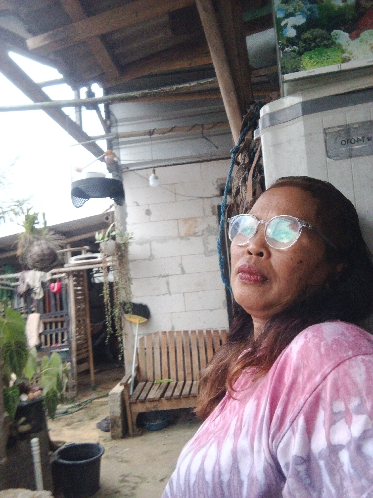 In an outdoor setting in Warungkiara, Indonesia, a woman wearing a pink tie-dye t-shirt and clear-rimmed glasses is pictured. She is leaning against a white structure, her gaze directed upwards and to the right. Her lips are slightly parted, and she appears to be in mid-conversation or contemplation. In the background, a wooden bench sits beside a brick wall, and various objects including plants, a bucket, and a broom are visible. The scene is covered by a wooden roof supported by beams. The overall environment suggests a domestic or semi-outdoor living space.