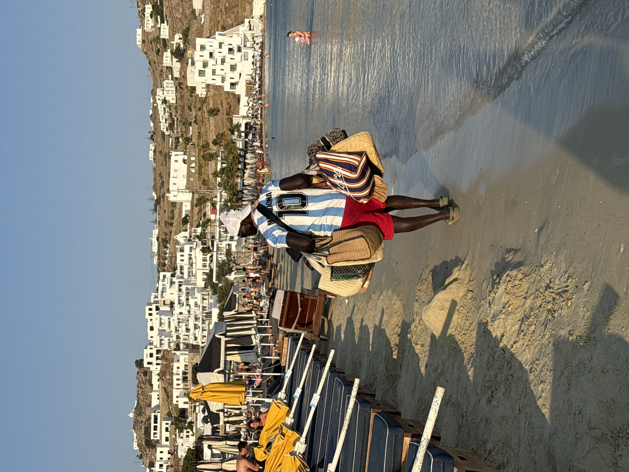 A man wearing a white cap and a blue and white striped soccer jersey with the name "MADONA" and the number "10" on the back walks along the sandy shore of Agios Ioannis Diakoftis, Greece. He is also wearing red shorts and tan sandals, and carries several large woven bags in his arms. The man is walking away from the viewer, towards the right side of the frame. The beach is lined with rows of blue lounge chairs with black and white striped canopies, and some yellow umbrellas. In the background, a hillside is covered with white buildings, typical of the Cycladic architecture found on the island. The sea is calm and the sky is clear blue. In the shallow water on the right side of the frame, a woman in a pink swimsuit is visible.
