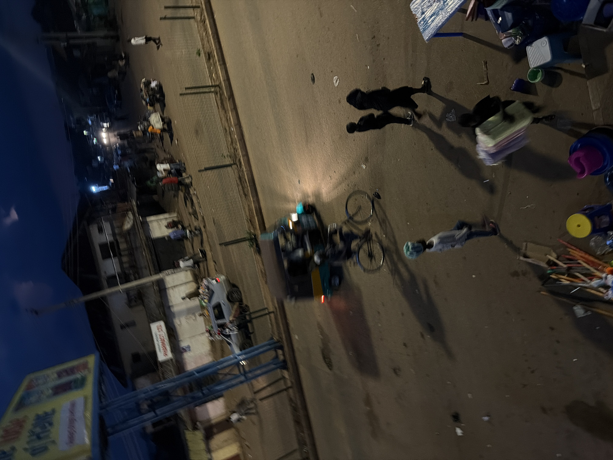 The image captures a street scene in Bukuru, Nigeria, at dusk or night. A blue and yellow auto-rickshaw is in motion, its headlights illuminating the road. In the foreground, a person on a bicycle is blurred, indicating movement. To the right, two individuals are walking away from the camera, their figures casting long shadows. Further down the street, a car appears to be stopped, with people standing nearby. Several buildings line the street, some appearing to be shops or residential structures. A prominent billboard with text in a foreign language stands on the left side of the image. The street is paved and appears to be a busy area, with scattered debris and trash visible on the ground. The overall lighting suggests artificial illumination from streetlights and vehicle headlights.