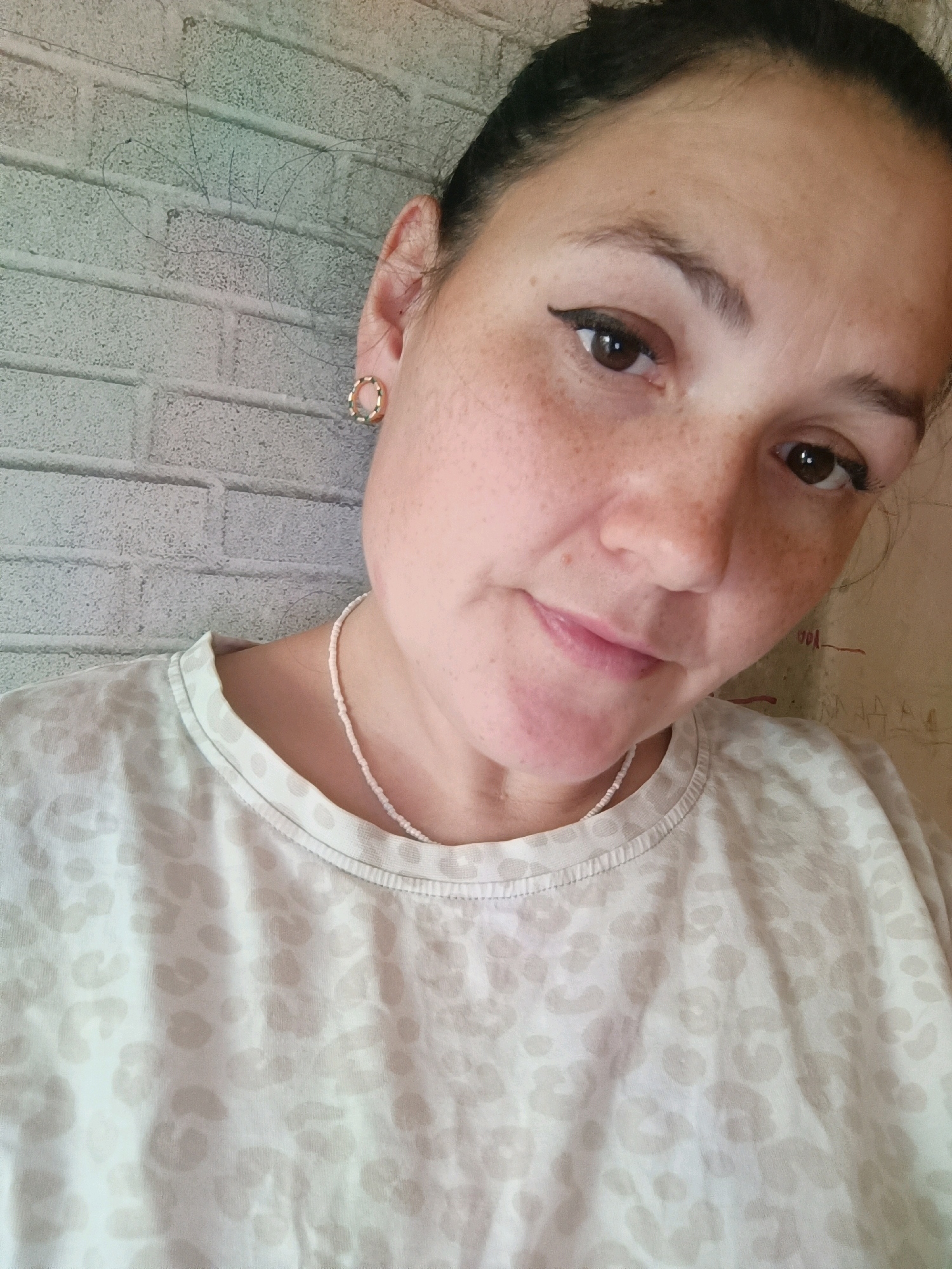 The image is a selfie of a woman with dark hair, brown eyes, and freckles. She is wearing a light-colored t-shirt with a subtle leopard print and a delicate beaded necklace. She has on small hoop earrings and her eyeliner is winged. The background appears to be a brick wall with a lighter shade of gray. The lighting suggests it may be daytime. The image was taken in Chaikovsky, Russia.