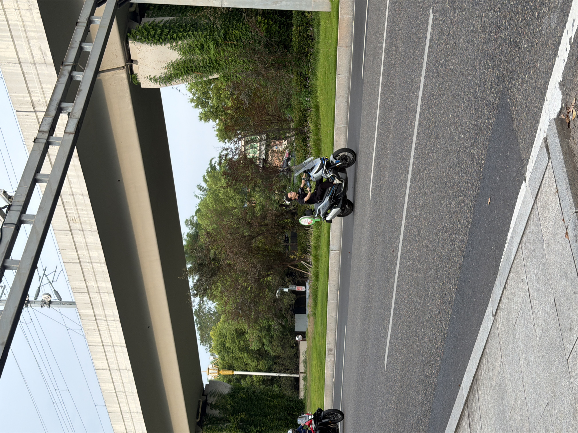 In Jiangjin, China, under an elevated highway structure, two individuals are riding motorcycles on a paved road. A person on a black and red motorcycle is in the left lane, partially obscured by the foreground. In the right lane, a person wearing a black short-sleeved shirt and dark pants is riding a silver and black scooter, with a clear windshield and a green circular object attached to the rear. The environment consists of green trees, grass, and a concrete sidewalk. The sky is overcast.