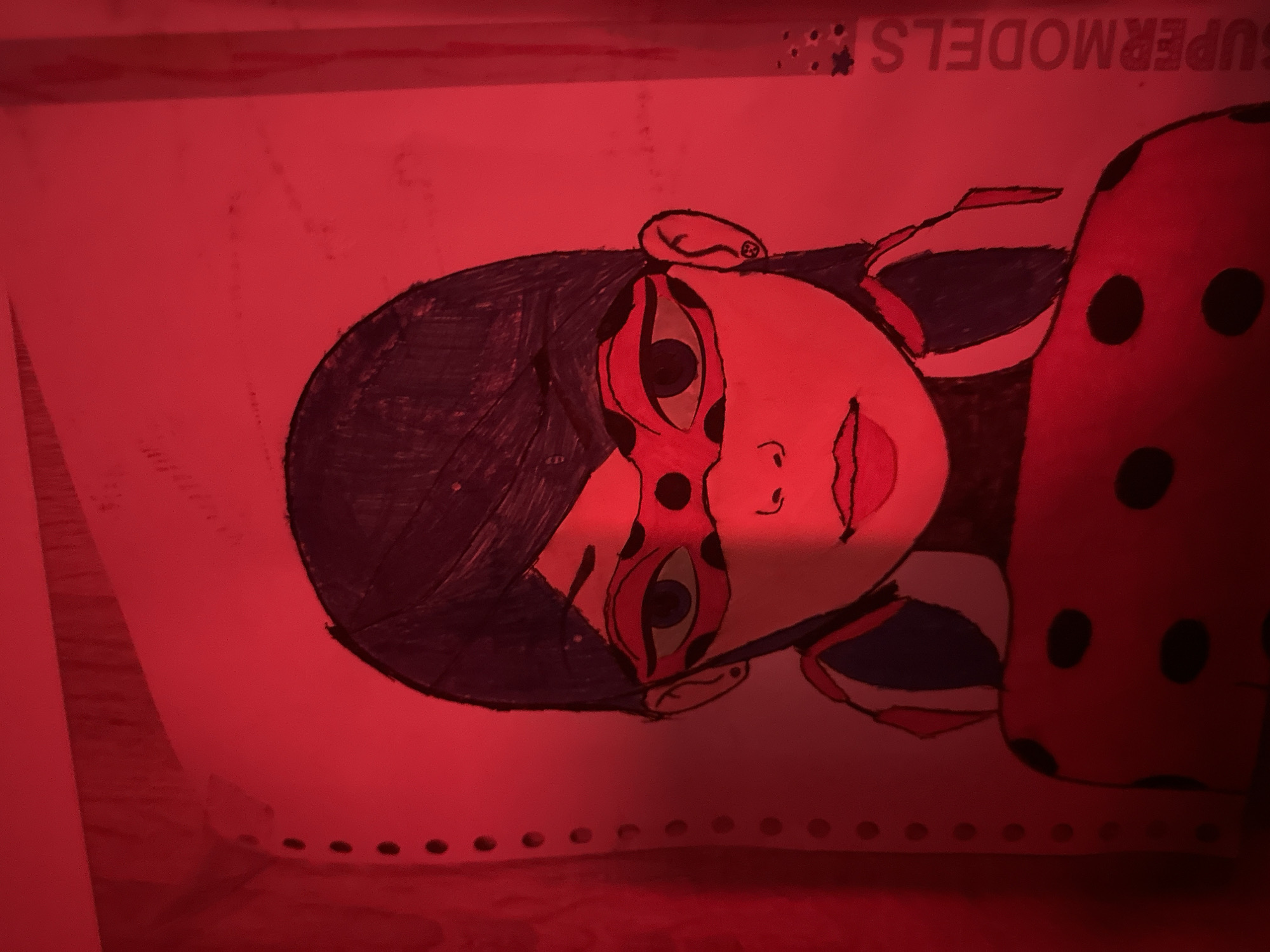 The image is a close-up shot of a drawing of a female character, likely from an animated series, wearing a red and black spotted outfit and a matching mask. The drawing is on lined paper with punched holes along the left side. The drawing is illuminated by a bright red light, casting a strong red hue over the entire image. The text "SUPERMODELS" is visible vertically along the right edge of the paper. The exact location of the drawing within the city of Unknown, Unknown cannot be determined from the provided image.