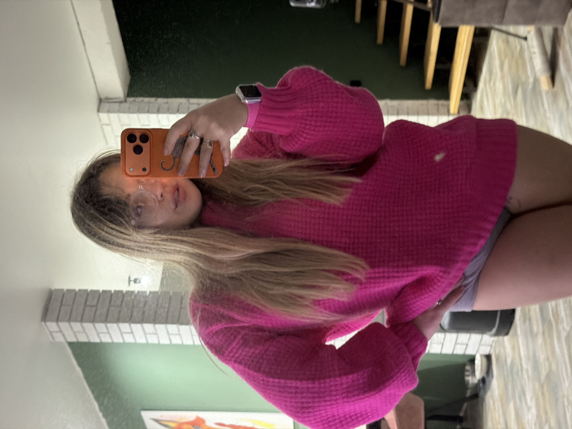 A woman is taking a selfie in a room. She is wearing a pink sweater, glasses, and an Apple Watch. She is holding an orange phone in her left hand. The room has green walls, a brick-like design, and a chair with wooden slats. The location is possibly in Большая Шильна, Russia.