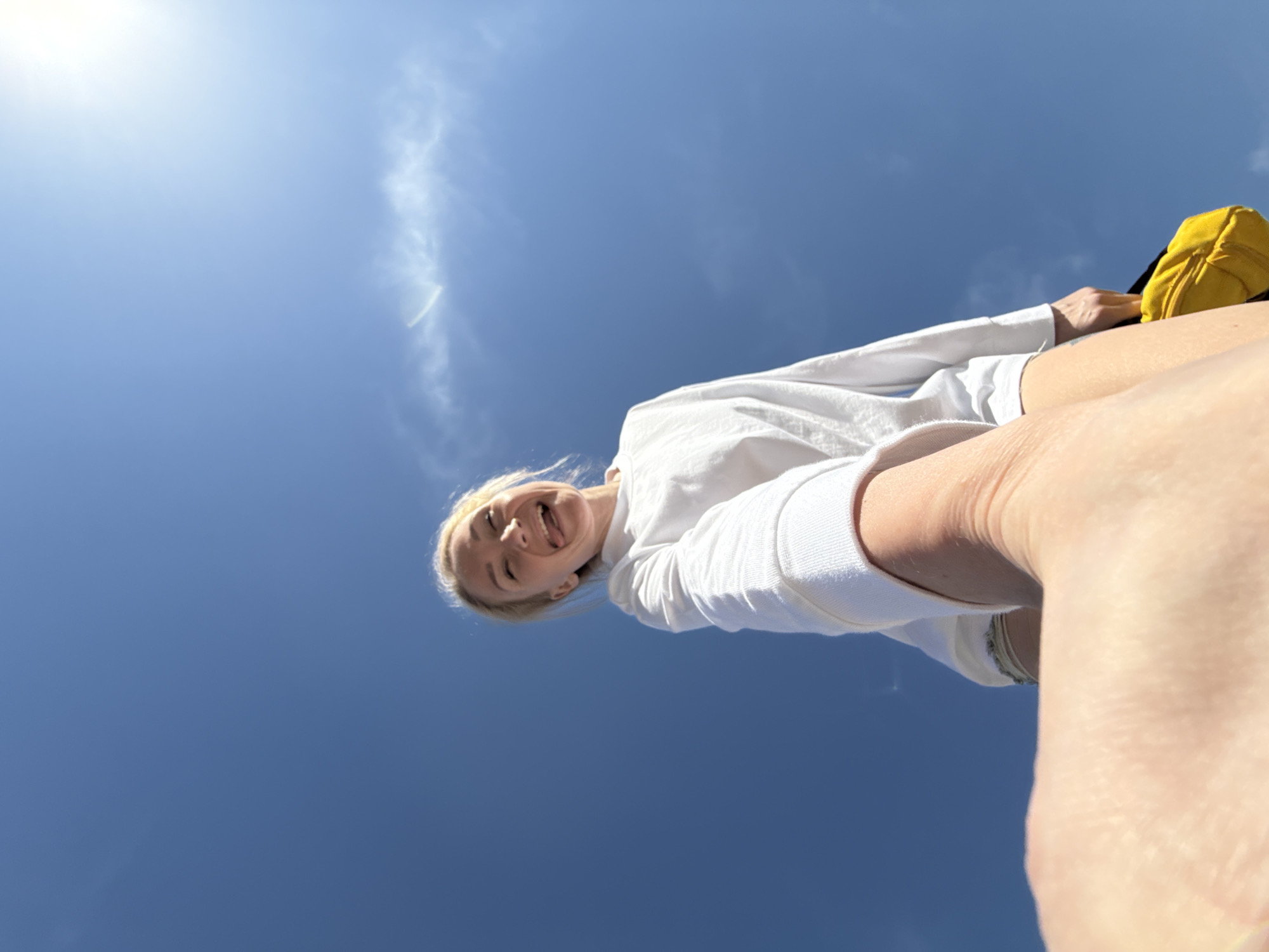 A low-angle shot looking up at a woman against a clear blue sky with a few wisps of white clouds. The woman is wearing a white long-sleeved shirt and appears to be sticking her tongue out playfully. A yellow backpack is partially visible in the top right corner of the frame. The location is impossible to tell based on the current image.