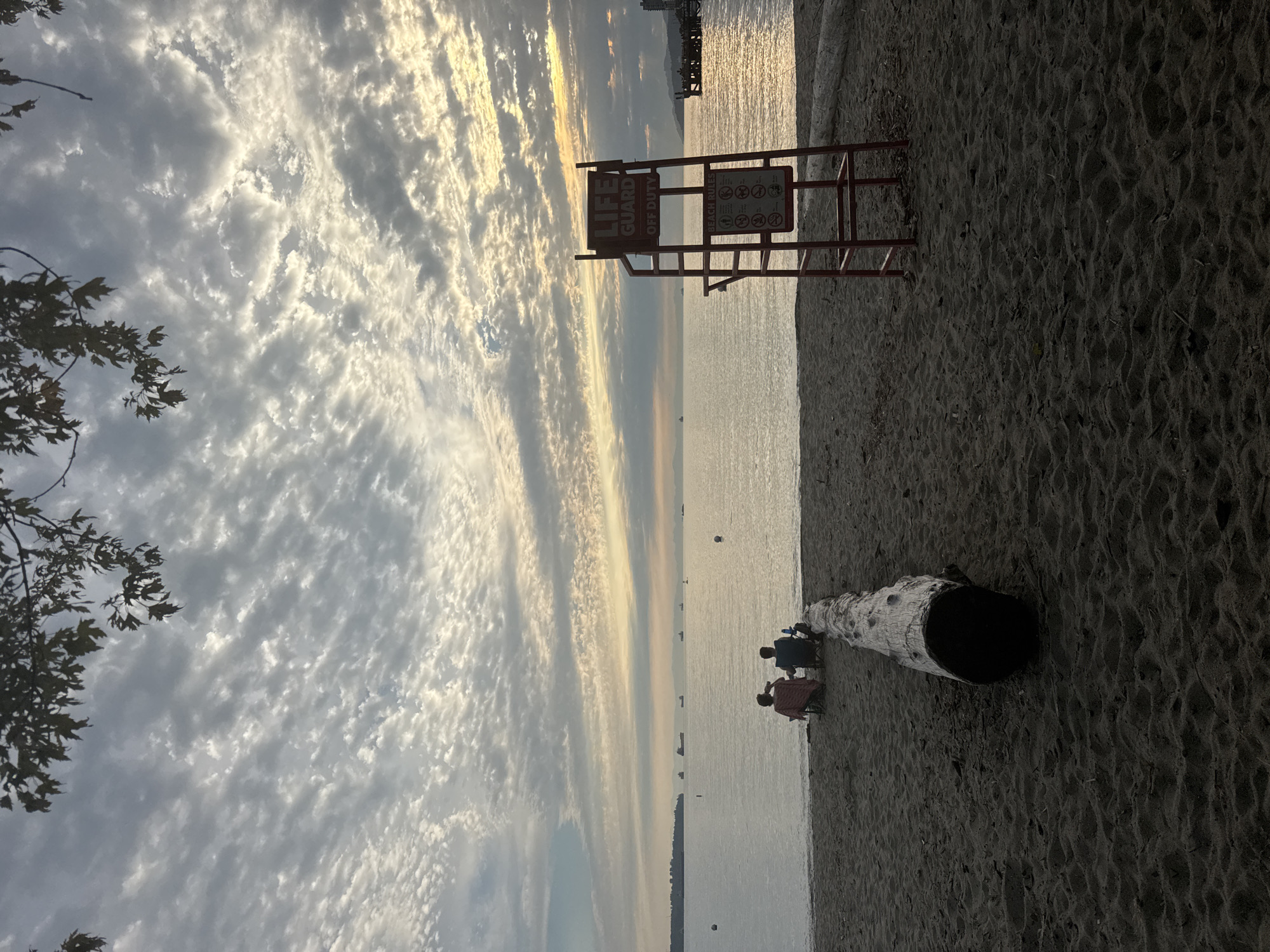 The image depicts a serene beach scene in West Vancouver, Canada, likely at sunset or sunrise given the soft lighting and atmospheric clouds. In the foreground, a large piece of driftwood rests on the dark sand. To the right, a red lifeguard stand is positioned, with a sign indicating "LIFE GUARD OFF DUTY" and "BEACH RULES." In the middle ground, two individuals are seated on beach chairs facing the water, one holding a drink. The calm ocean stretches out to the horizon, dotted with several large ships. The sky above is filled with a dense, textured pattern of white and gray clouds, with a warmer glow of light visible on the right side. In the upper portion of the image, tree branches with leaves hang into the frame from the top.