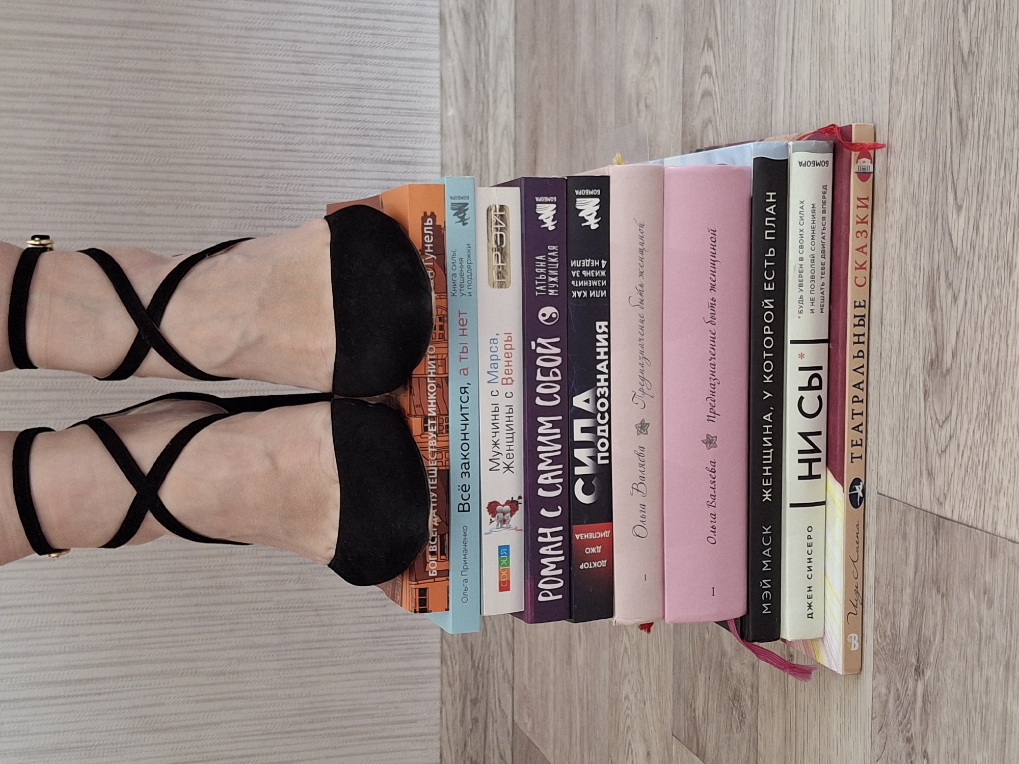 Here is a description of the image.
The image shows a top-down view of a row of ten books stacked vertically on a light-colored wooden floor. The books appear to be mostly self-help or relationship-oriented titles, with Russian text on their spines. A pair of women's black heeled sandals with criss-cross straps, are positioned with the heels obscuring the spines of the first few books. The focus is on the composition of the shoes and the books, with the floor providing a neutral background. The location is unknown.