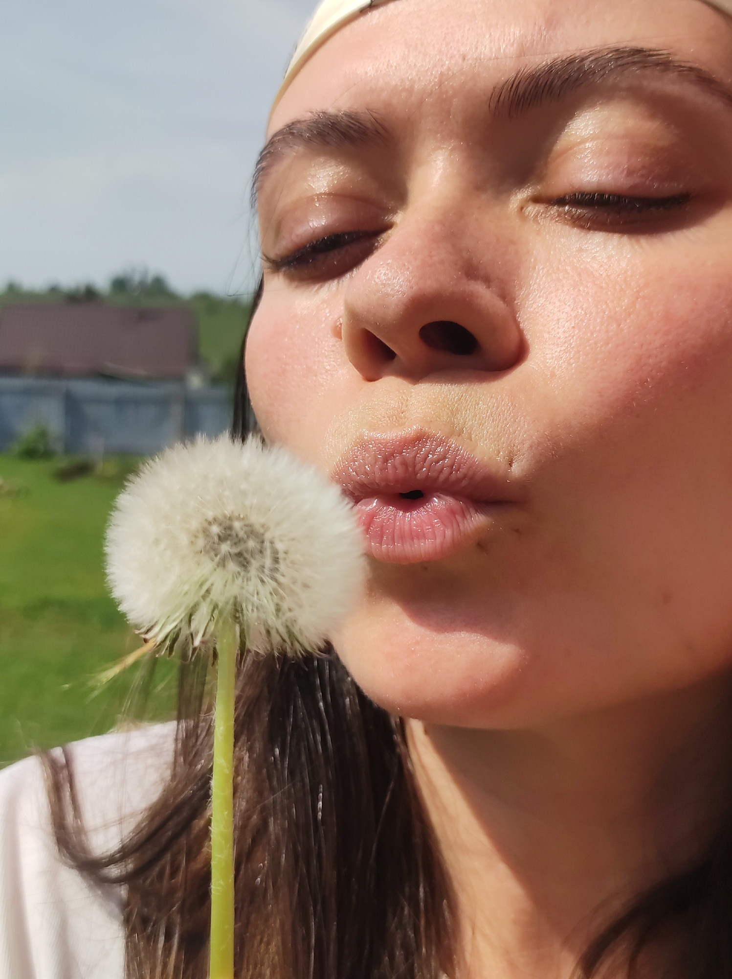 A close-up captures a fair-skinned woman, likely in her early twenties, with her eyes closed, pursing her lips toward a dandelion. She wears a light-colored headband and a white top. Her dark hair falls down her shoulders. The dandelion, prominently positioned, is in sharp focus. The background includes a blurred building with a brown roof and a gray fence, suggesting a rural or suburban environment. The location is Фоки, Russia, as stated in the prompt. The environment appears bright and sunny.