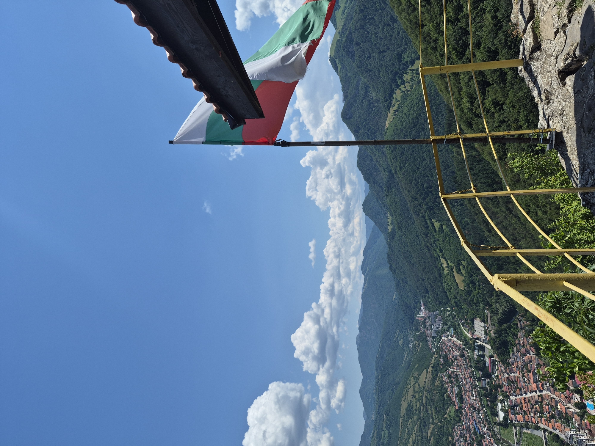 The image depicts a scenic overlook in the city of Teteven, Bulgaria, on a sunny day with a bright blue sky dotted with fluffy white clouds. In the foreground, a yellow metal railing runs along the edge of a rocky outcrop, providing a safety barrier. A flagpole with the Bulgarian flag, featuring white, green, and red horizontal stripes, is prominently displayed on the right side of the frame. The flag is partially obscured by a section of a tiled roof structure extending from the upper right corner.
Beyond the railing and flag, a panoramic view unfolds of the Teteven valley. Lush green, densely forested mountains stretch into the distance, their slopes creating a dramatic, undulating landscape. Nestled within the valley is the city of Teteven, characterized by a dense cluster of buildings with terracotta roofs, interspersed with streets and some larger structures that appear to be municipal or educational buildings. A river or road can be seen winding through the urban area. The overall impression is one of natural beauty and a well-preserved historical town situated within a mountainous region. There are no people visible in the image.