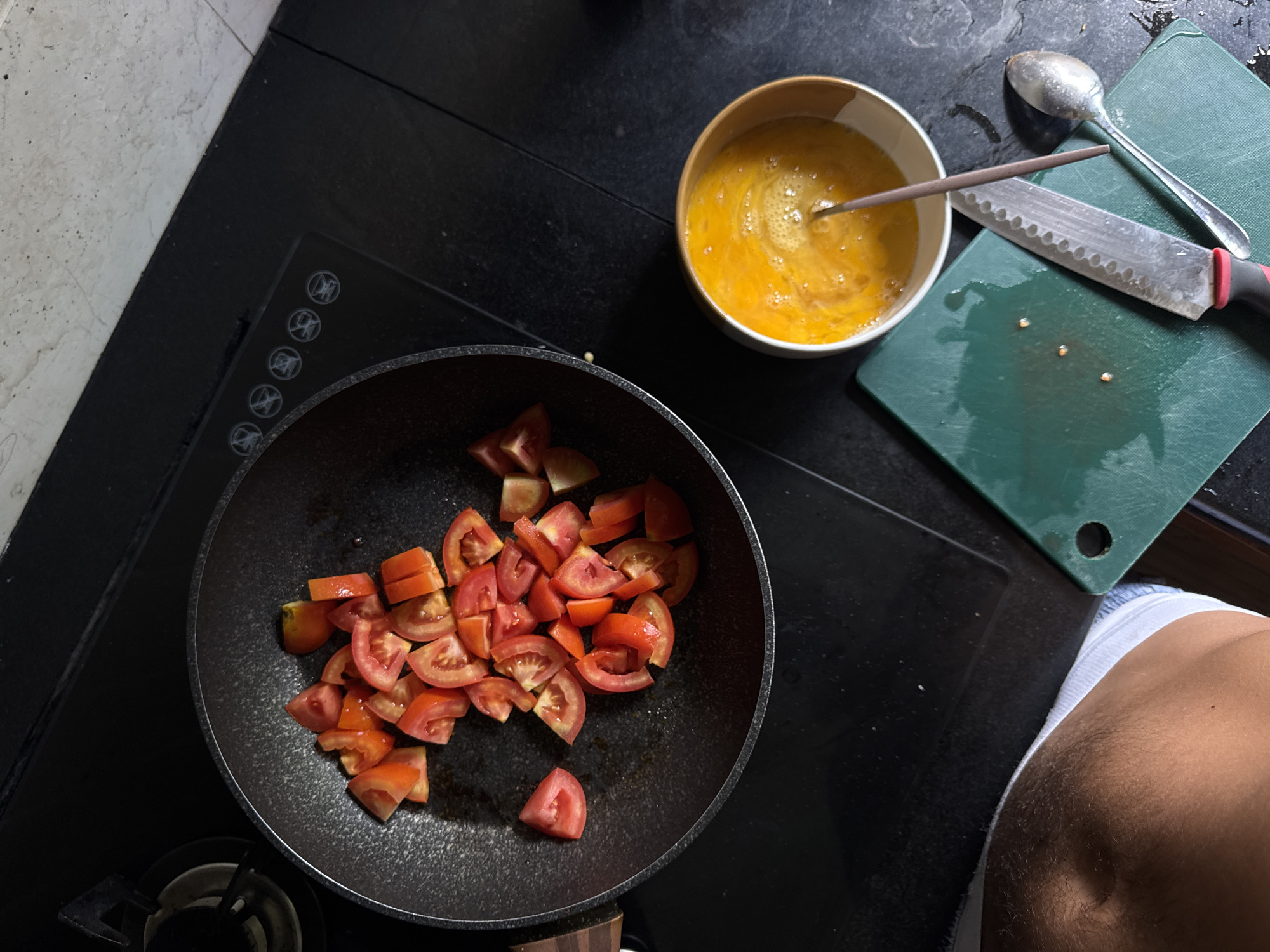 An overhead shot shows a person's legs and midriff, wearing white underwear, to the left of the frame. The scene appears to be a kitchen with a black induction stove. On the stove, a black frying pan contains chopped tomatoes. To the right of the pan, a white bowl holds beaten eggs. A green cutting board sits below the bowl, with a knife and spoon on top of it. The background is a dark countertop and a light-colored tiled wall. The image is likely taken in Kuta Selatan, Indonesia, a popular tourist destination known for its beaches and culinary scene.