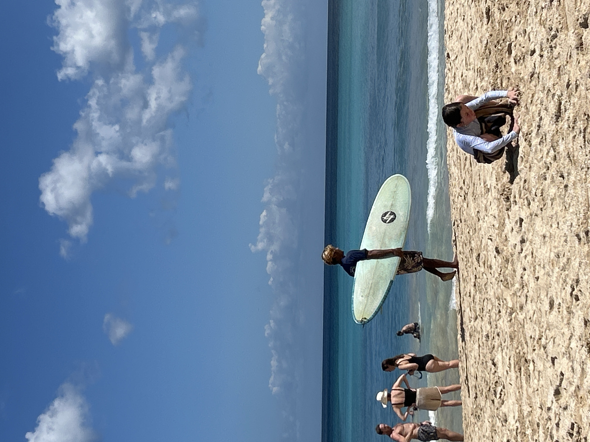 This image depicts a beach scene in Kuta Selatan, Indonesia, under a bright, partly cloudy sky. The foreground is dominated by sandy beach where a young person in a long-sleeved rash guard and swim trunks is crouched down, seemingly examining something on the sand.
Further back, a man with blonde hair, wearing a dark shirt and patterned shorts, carries a light-colored surfboard under his right arm. He is walking towards the water. A group of three individuals are closer to the water's edge. To the left, a man in patterned shorts stands, facing away from the camera. Next to him, a woman in a wide-brimmed hat and a black swimsuit is looking towards the man with the surfboard. A third woman, in a black swimsuit, stands slightly behind and to the right of the woman with the hat, also facing the water.
In the middle ground, the turquoise and dark blue ocean stretches out, with gentle waves breaking near the shore. A person is partially visible in the water, sitting in the shallows. The horizon is clearly defined where the water meets the sky, which is a vibrant blue with scattered white cumulus clouds. The sand appears to be a light tan color with visible textures and small undulations.