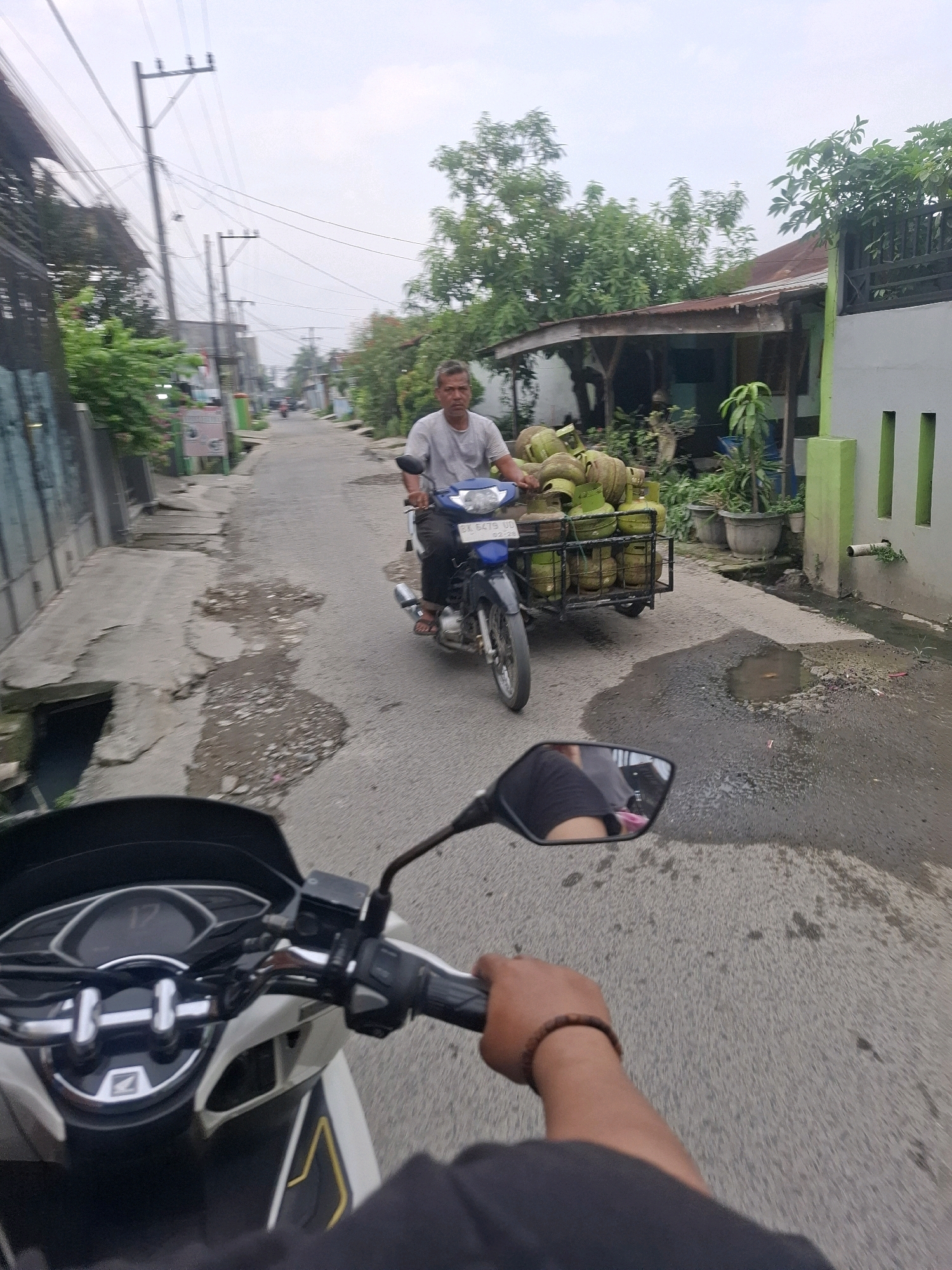 The image depicts a street scene in Tembung, Indonesia, captured from the perspective of someone riding a motorcycle. In the middle ground, a man is riding a blue motorcycle with a trailer attached, carrying a load of green gas cylinders. He is wearing a grey t-shirt and dark pants. The street is paved and appears to have some potholes and puddles. On either side of the street are buildings, including a house with a reddish-brown roof and a grey wall with green accents. There are trees and vegetation visible. In the foreground, the handlebars and dashboard of the motorcycle the viewer is riding are visible. The sky is overcast.