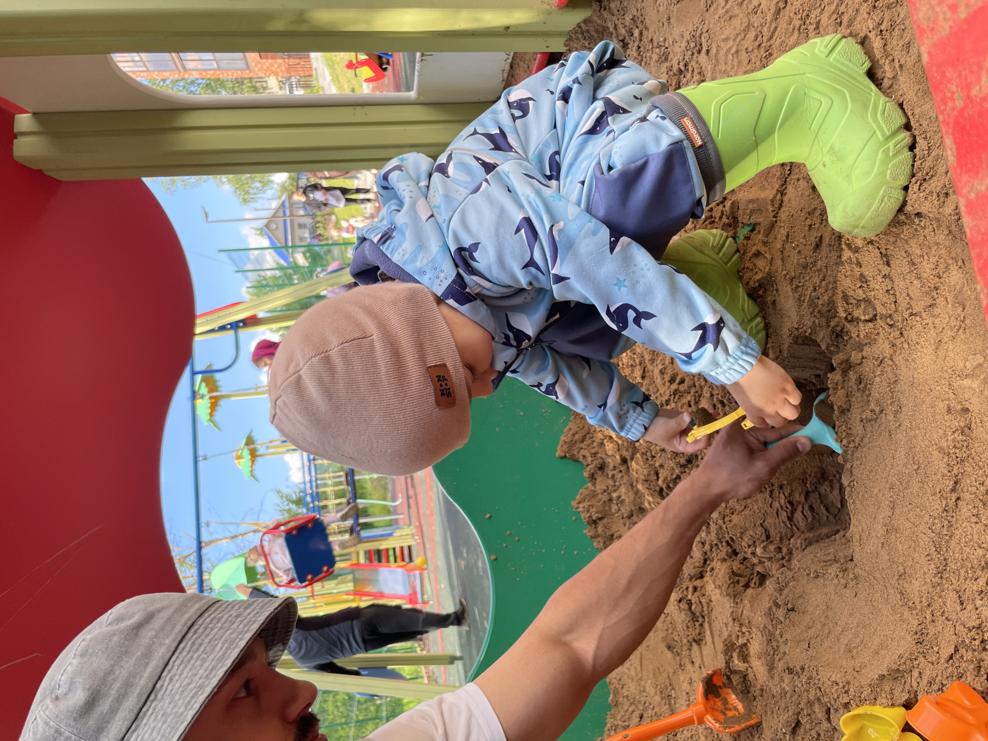 The image depicts a vibrant outdoor scene at a playground, focusing on a sandpit activity. A young child, likely a toddler, is the central subject, dressed in a light blue jumpsuit featuring dark blue whale and star patterns. The child also wears a light brown ribbed beanie with "NCS" and a small logo visible, and bright green rubber boots, one of which clearly shows "Nordman" on its grey cuff. The child is bent over, actively playing in the brown sand, holding a yellow sand tool.
An adult's muscular arm, with darker skin, is seen reaching into the sandpit from the foreground, holding a light blue sand shovel. This adult, whose face is partially visible in the bottom-left corner looking upwards, wears a grey bucket hat and a white t-shirt. The adult appears to be interacting with the child, possibly assisting or playing alongside them. Other colorful sand toys, including an orange shovel and yellow and orange molds, are scattered in the sand.
The immediate surroundings include red and light green sections of a playground structure, framing the sandpit. In the background, under a clear blue sky with some white clouds, other playground equipment, green trees, and residential buildings can be seen. Another adult figure is visible further in the background on the playground surface. The scene suggests a sunny daytime setting, filled with playful engagement between the child and adult in a typical playground environment located in Izhevsk, Russia.