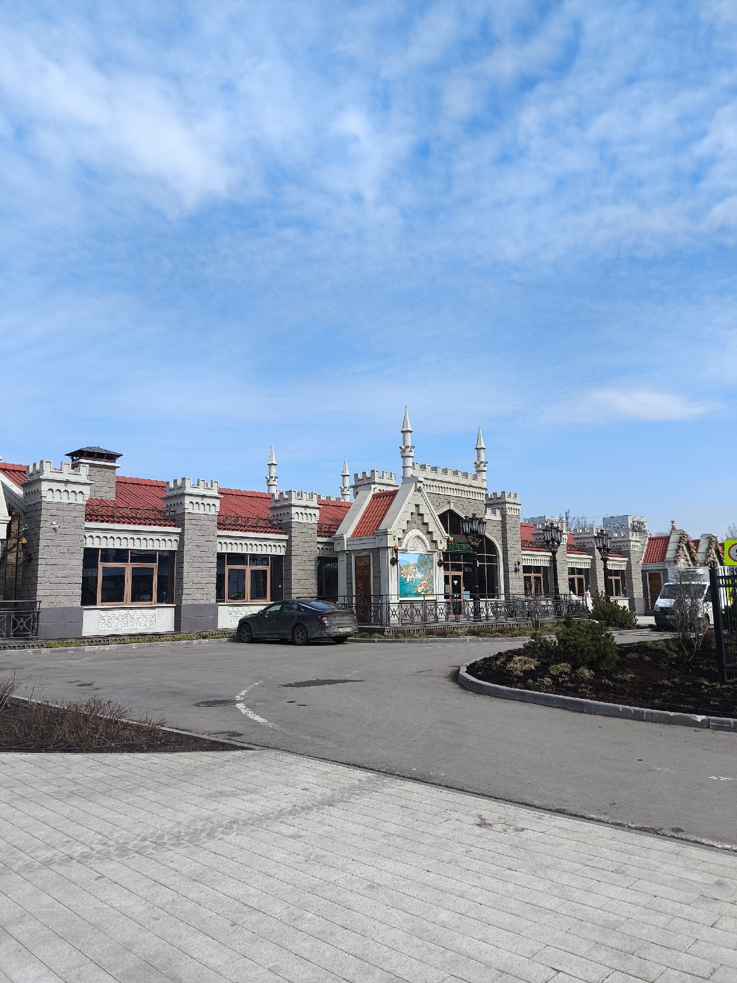 The image shows a building with castle-like architecture in Moscow, Russia. The building has a grey stone facade, red roof, and decorative turrets. A grey sedan is parked in front. In the foreground, there's a paved area and a circular landscaped island with small bushes. The sky is blue with scattered clouds. A street sign is visible on the right.