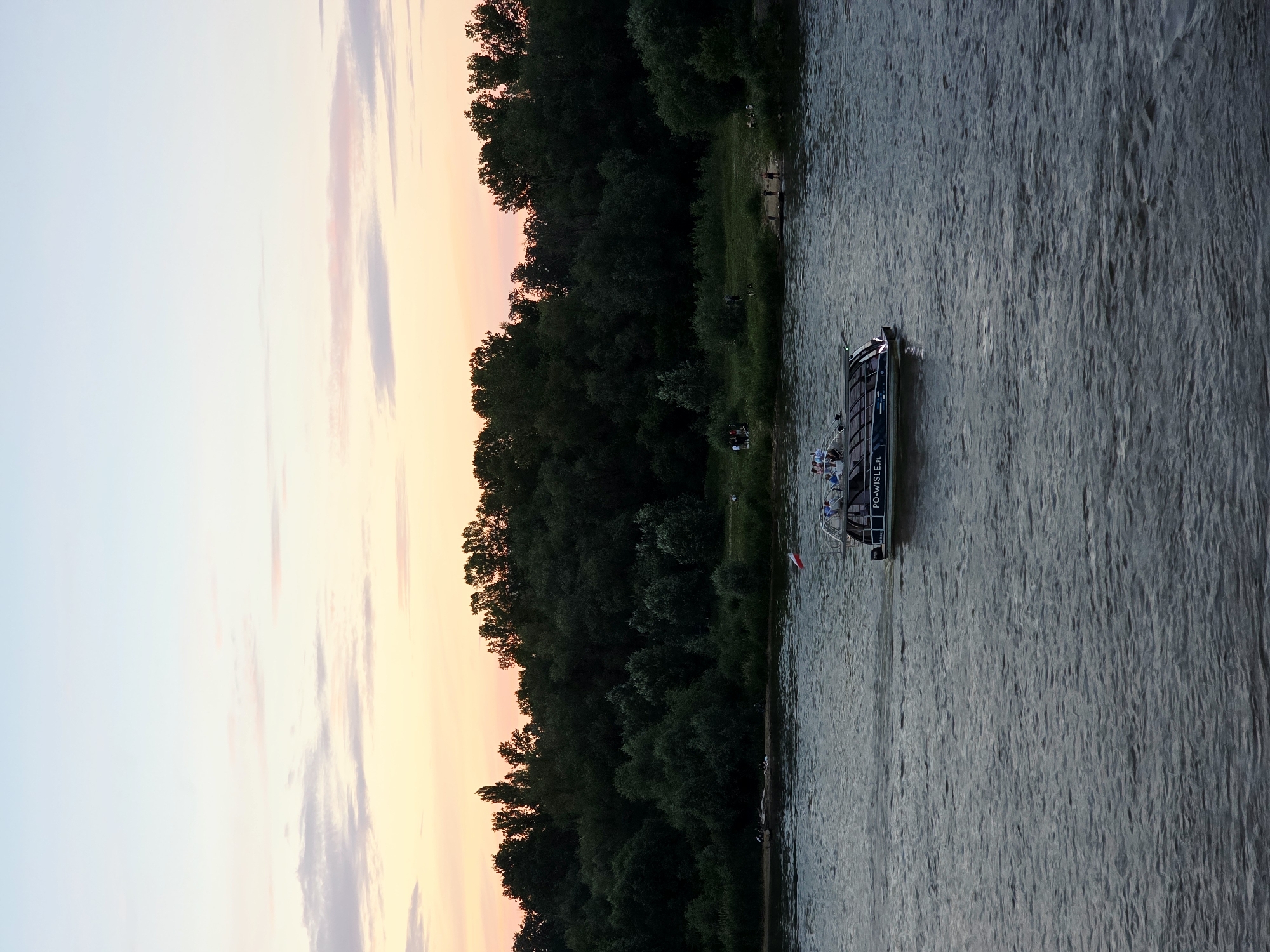 This is an aerial, medium shot of a boat on the Vistula River in Warsaw, Poland, at dusk. The boat is dark with a glass roof and has the text "PO-WISLE.PL" on its side. There are people on the boat, and a Polish flag is flying from its stern. The river is dark and rippling. In the background, a dense line of trees forms the riverbank, with some people visible on the sandy shore in the distance. The sky above is a soft gradient of pale blue, pink, and orange, indicating the setting sun. The overall impression is one of peaceful evening activity by the river.