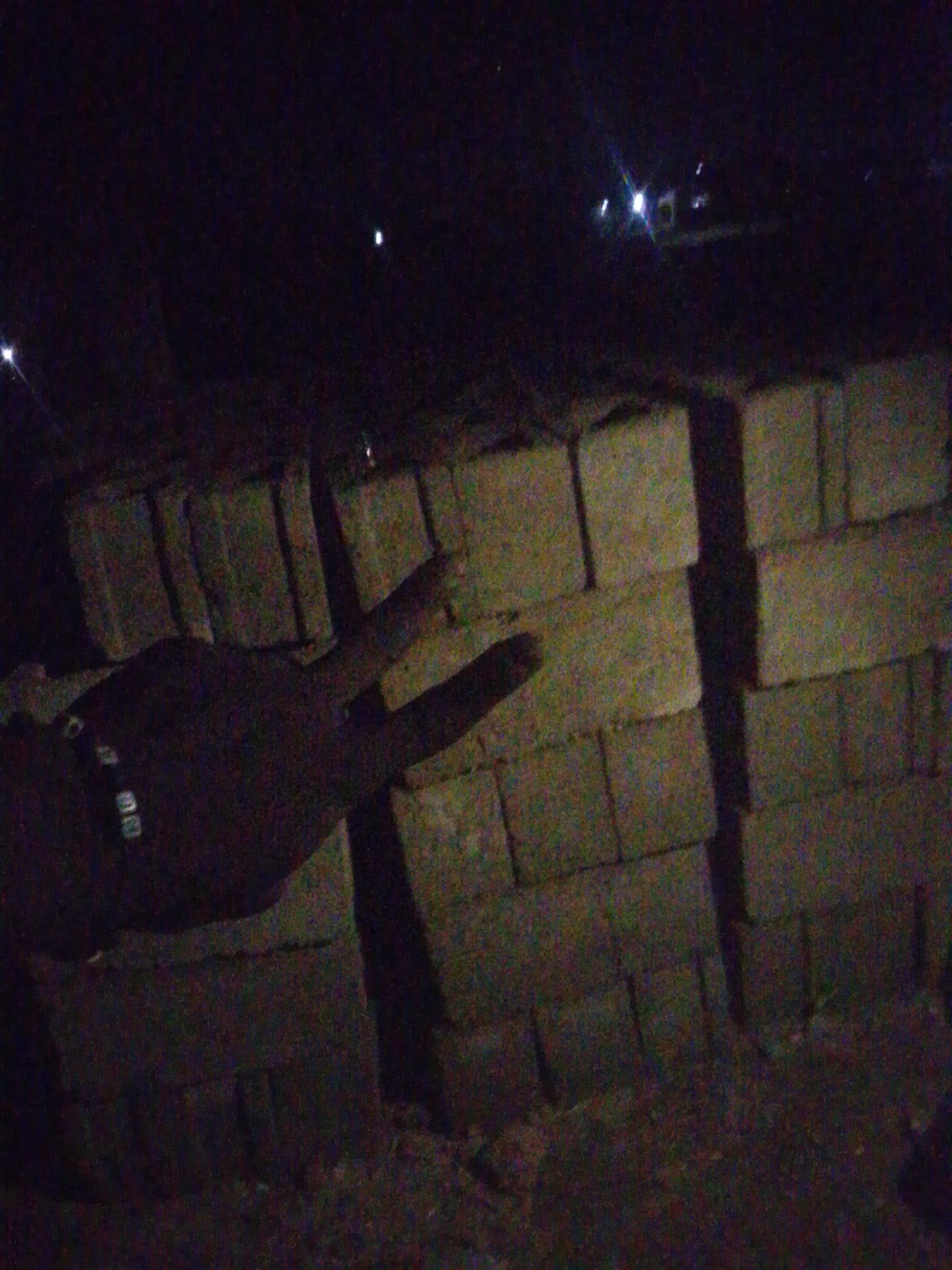 A low-light, high-noise photograph depicts an outdoor scene at night in the city of Unknown, Unknown. In the left foreground, a human hand, possibly dark-skinned or heavily shadowed, extends into the frame from the bottom left, displaying the index and middle fingers upright in a "V" gesture, commonly known as a peace sign. A band with light-colored, rectangular elements is visible on the wrist. Behind the hand, and extending across the midground, are multiple stacks of concrete blocks or bricks, forming irregular piles or incomplete walls. The ground beneath is dark and indistinct. The background is predominantly dark, with several distinct, distant artificial light sources visible, some exhibiting lens flare, suggesting an urban or semi-urban environment.