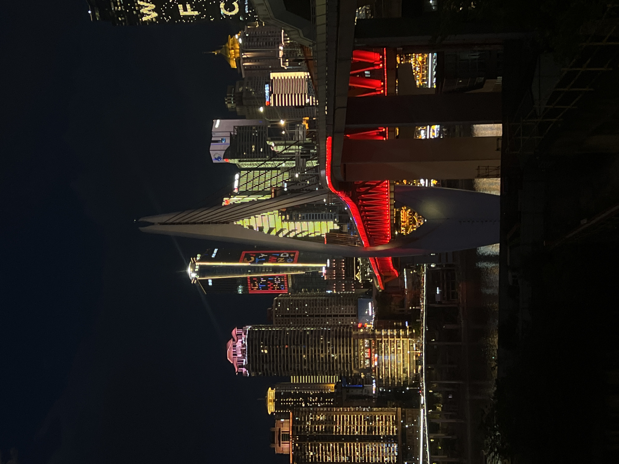 The image shows a nighttime cityscape with a suspension bridge illuminated in red as a prominent feature in the foreground. Behind the bridge, a dense collection of illuminated skyscrapers of varying heights and architectural styles form the urban skyline. The buildings are adorned with a variety of lights, including vertical bands of green, yellow, and pink, as well as horizontal patterns. Some buildings have distinct illuminated tops. A river is visible at the bottom of the frame, reflecting the city lights. The sky is dark, indicating it is night. The bridge's structure is visible with its cables and a central tower, all contributing to the dynamic night-time ambiance. The specific location of 江北城街道, China, implies this is a scene from Chongqing, a major city in China known for its dramatic riverside architecture and impressive bridges.