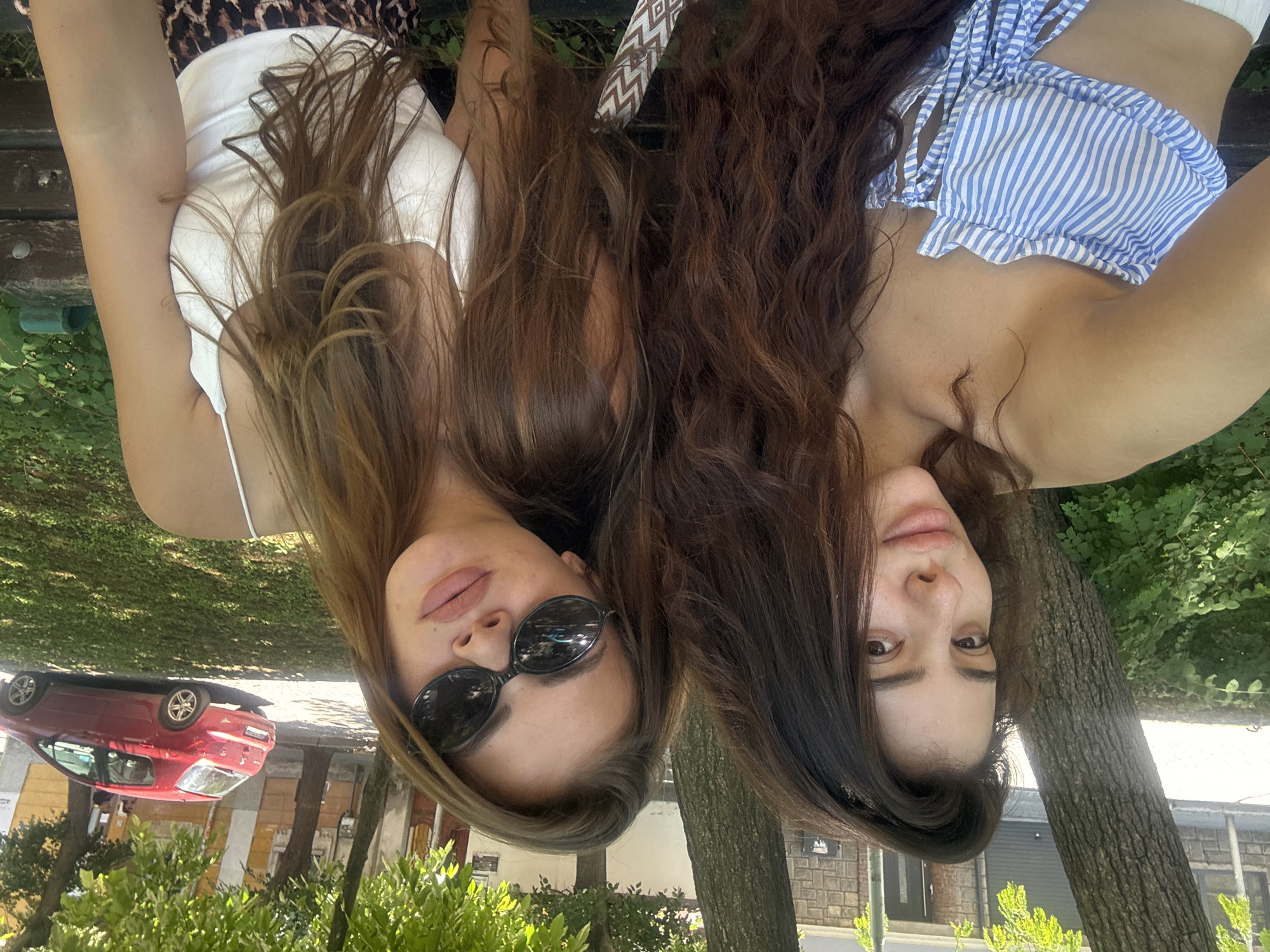 Two young women are posing for a selfie on a wooden bench in an outdoor setting, possibly a park or a quiet street in Plovdiv, Bulgaria. The woman on the left has long, dark, curly hair and is wearing a blue and white striped strapless top. She is looking directly at the camera with a neutral expression. The woman on the right has long, light brown hair and is wearing a white tank top and large dark sunglasses. She is also looking towards the camera, with her head tilted slightly. Her lips are slightly parted.
In the background, there are trees with lush green foliage, casting shadows on the ground. Beyond the trees, a red car is parked on the street, and some buildings with yellow accents are visible. The overall impression is a casual, sunny day captured in a candid moment. The image appears to be taken during daylight hours, given the bright lighting and clear visibility of the surroundings. The presence of trees and buildings suggests an urban or semi-urban environment.