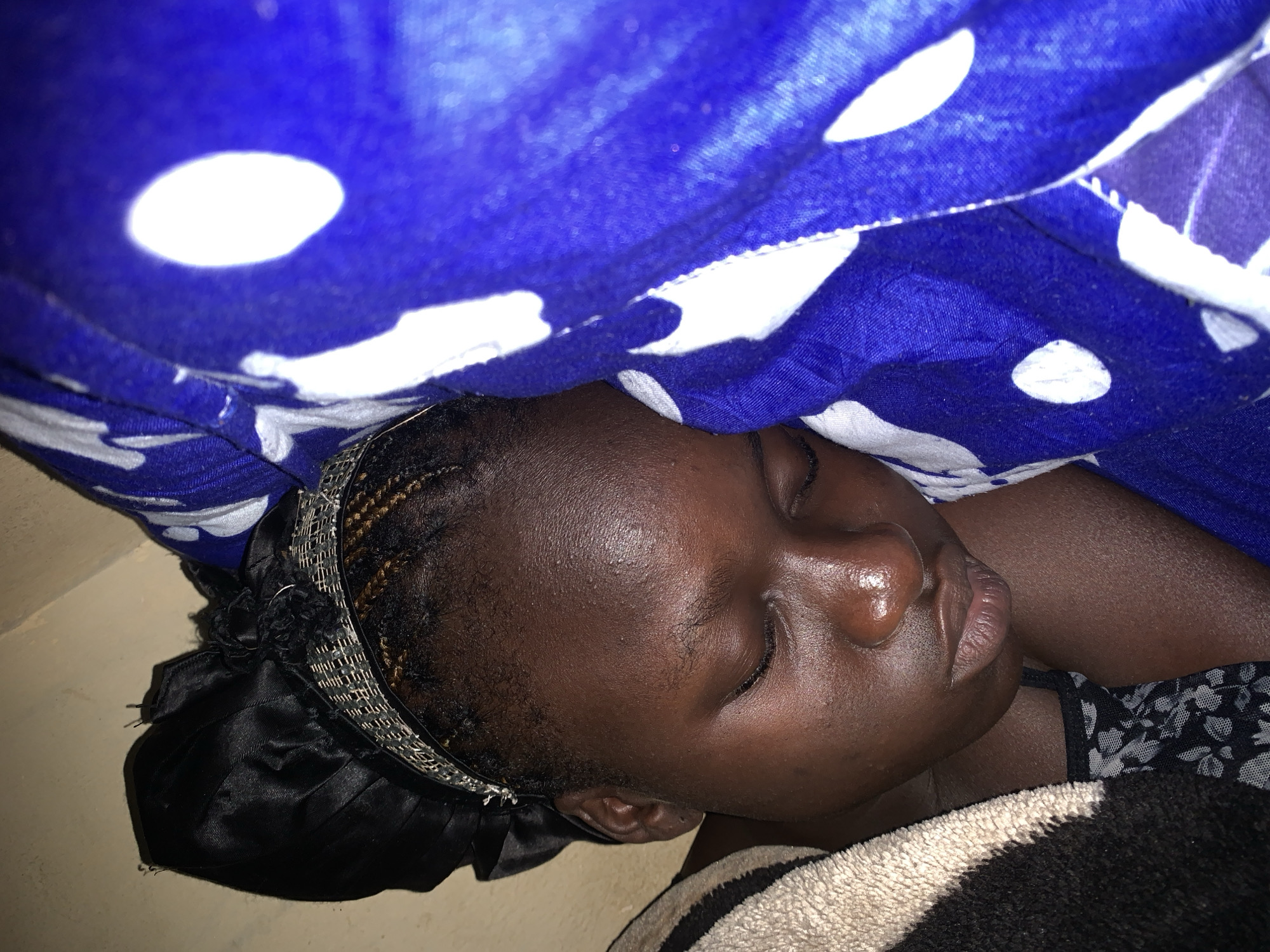 A close-up, overhead shot shows a dark-skinned person sleeping, positioned on their left side. The subject is wearing a black satin bonnet, secured by a metallic headband with a textured pattern. Their eyes are closed, and their head rests on a beige and black striped blanket. To the right of the subject, a blue fabric with white polka dots is visible, appearing to be bedding or a blanket. The overall lighting is dim, with a slight yellow cast, suggesting an indoor setting. The background is an indistinct, light-colored wall. The image does not provide any specific details to determine the location within a city.