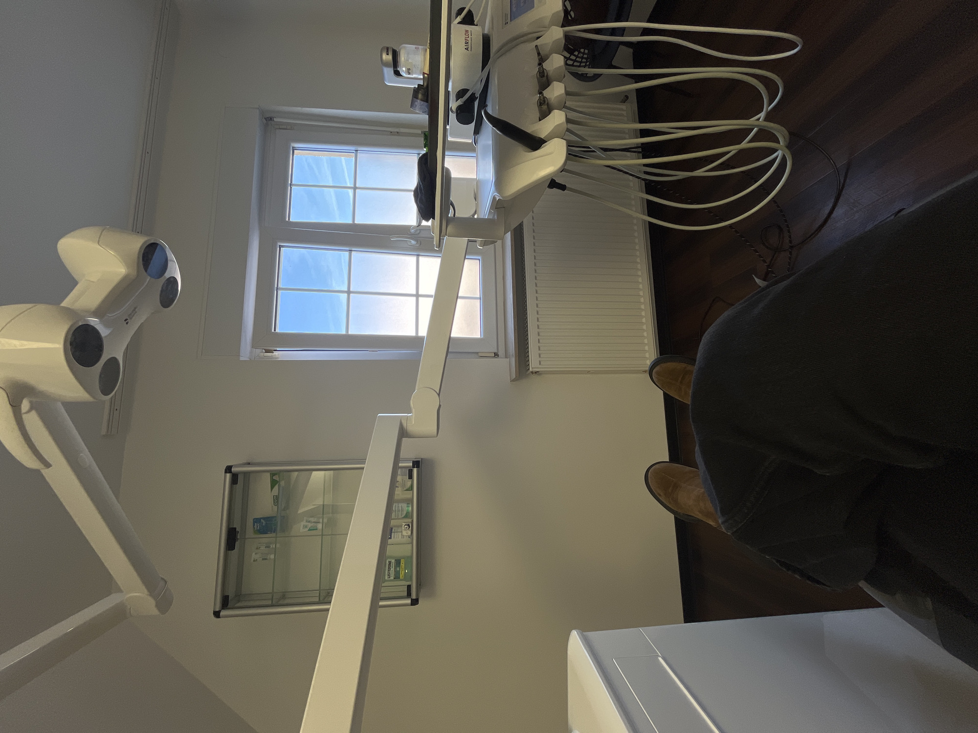 The image depicts the interior of a dental examination room located in Monsheim, Germany, from a first-person perspective, looking forward from a seated position. The lower legs of a person wearing dark trousers and brown shoes are visible in the foreground. The room features white walls and a dark wooden floor. A large window, segmented into multiple panes by white frames, dominates the right side of the frame, admitting bright natural light and revealing a blue sky with faint clouds. A white radiator is positioned directly beneath the window.
To the left, a silver-framed glass display cabinet is mounted on the wall, containing various dental hygiene products, including bottles of Listerine mouthwash and packaging for GUM brand items. Above, a white, articulated dental examination light fixture with multiple lamp heads is suspended from the ceiling. A long, white, articulated arm extends into the mid-ground, likely part of the dental chair or unit's support structure.
To the right, a multi-component white dental unit is situated near the window. It features multiple white hoses with black tips hanging from its side, typical of dental instruments. On the unit's top surface, a dark folded cloth, a small silver object, and a white bottle labeled "NFS" are present. The "AIRFLOW" brand logo is visible on the main body of the dental unit. No other individuals or active procedures are depicted.