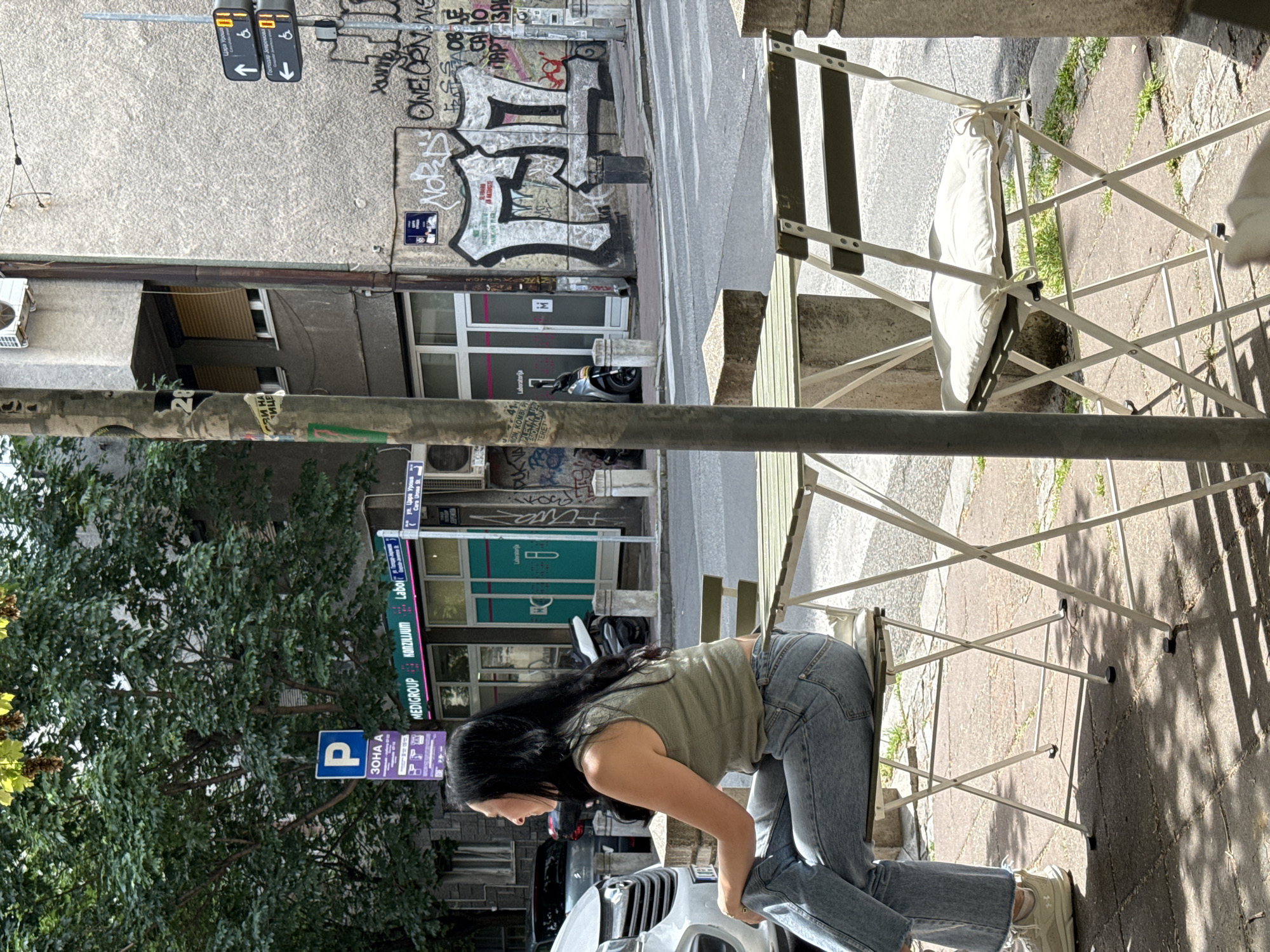 A woman in jeans and a tank top is bending over next to a folding chair in Belgrade, Serbia. Graffiti covers the building in the background and a street sign points toward "Gospodar Jevr." Additionally, there's a parking sign labeled "ZONA A", the "MEDIGROUP KONZILIJUM Labo" sign, and a laboratory sign, along with a pedestrian crossing sign on the building.