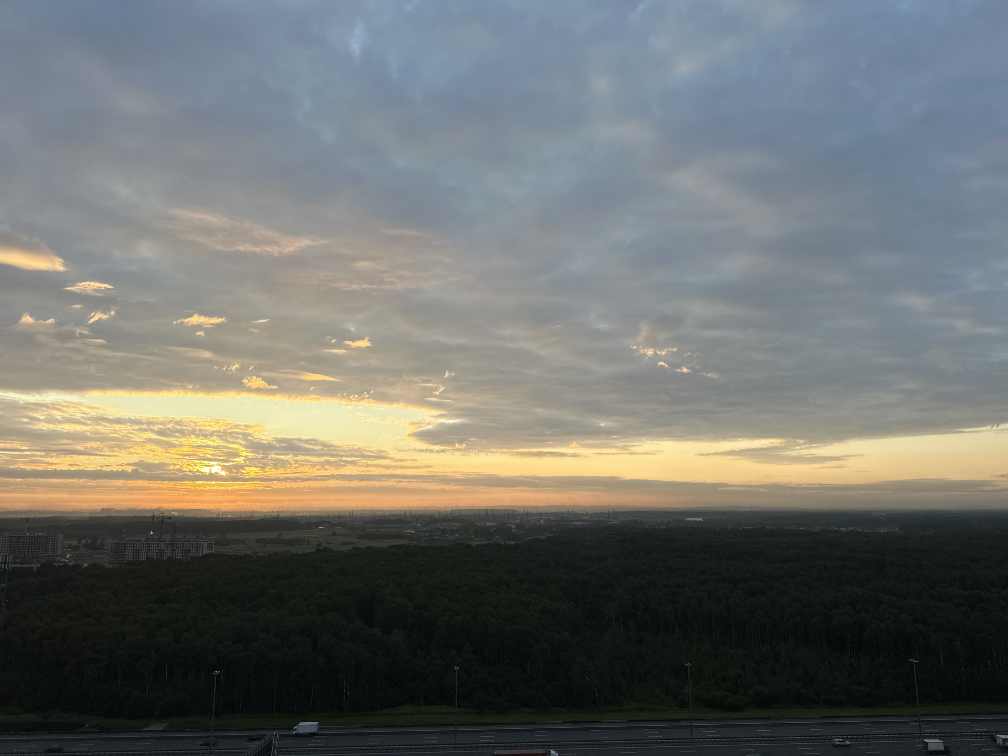 The image is a wide-angle shot capturing a sunrise over a suburban landscape, likely Kudrovo, Russia. The sky is filled with a mix of gray and white clouds, with patches of orange and yellow sunlight breaking through on the left horizon, indicating the early morning. Below the sky, a vast expanse of dark green forest dominates the middle ground. In the far distance, a hazy cityscape is visible with industrial structures and what appear to be tall residential buildings under construction on the left. In the foreground, a multi-lane highway runs horizontally across the frame, with a few vehicles, including cars and trucks, on it. Streetlights are visible along the side of the highway. The overall scene depicts a transition from night to day, with the natural environment of the forest contrasting with the developing urban infrastructure.
