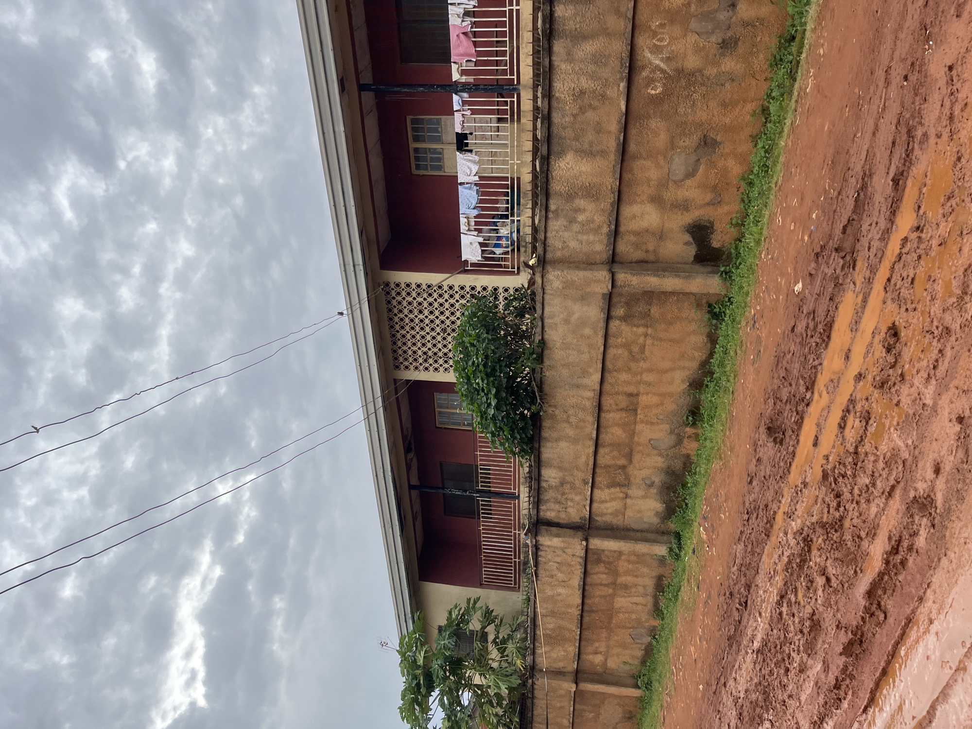 A low-angle, medium shot captures a section of a residential building and a muddy dirt road in Bukuru, Nigeria. The building, painted in a muted yellow and a deep red, features a covered porch with a white metal railing where laundry is hung out to dry. A decorative perforated concrete screen is visible on the left side of the porch. A substantial, weathered concrete wall, approximately waist-high, runs horizontally across the foreground, separating the road from the building. Beyond the wall, a lush green bush is situated to the left of the porch, and another bushy plant is visible on the right. The sky above is overcast with dense, grey clouds. In the immediate foreground, a rutted dirt road is covered in thick, reddish-brown mud and puddles, indicating recent rainfall. Two dark electrical wires stretch across the upper portion of the frame. No people are visible in the image.
