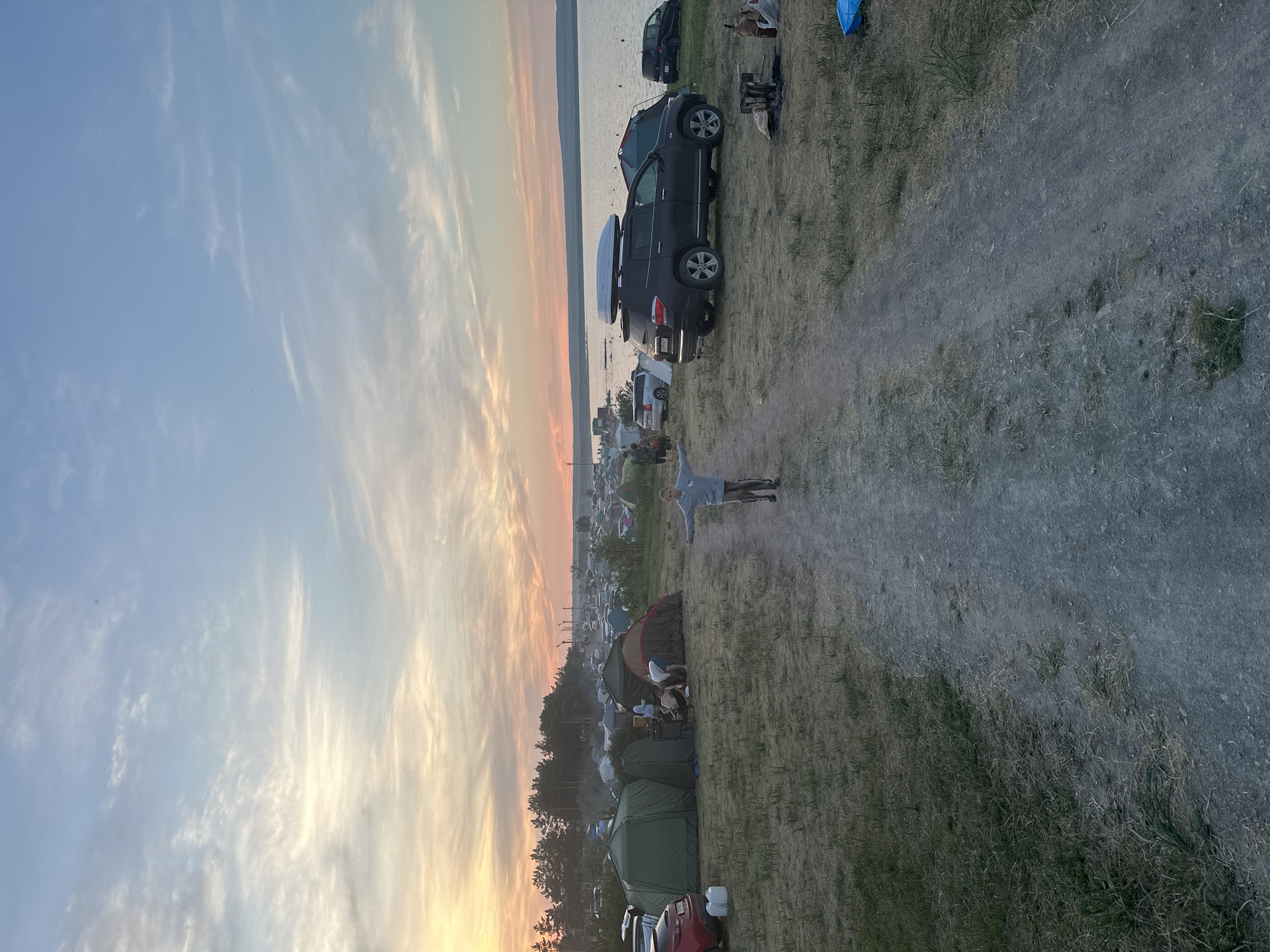 A wide-angle, eye-level shot captures a bustling campsite at dusk, likely near the city of Zhemchuzhny, Russia. The sky above is a canvas of soft blues and oranges, streaked with wispy clouds, suggesting the sun is either rising or setting over the adjacent body of water. A sandy, unpaved path runs down the center of the frame, leading the viewer towards a large gathering of tents and vehicles.
In the foreground, a young person stands with their arms outstretched, facing the viewer. They are wearing a light gray hooded sweatshirt and striped pants, and appear to be a child or young teenager. The path they stand on is well-trodden, indicating frequent use by campers.
Scattered across the grassy terrain are numerous tents of various sizes and colors, with some exhibiting signs of recent activity. Several vehicles are parked among the tents, including a dark SUV with a white roof box on top, a red car, and a black car. A small campfire pit, containing burnt wood, is visible on the right side of the path.
In the background, a dense line of trees forms a dark silhouette against the horizon. Beyond the trees, more tents and structures are visible, suggesting the campsite extends for a considerable distance. The calm water of a large lake or reservoir borders the campsite on the right side of the frame. The overall atmosphere is one of a lively outdoor gathering, possibly a festival or a large camping event.
