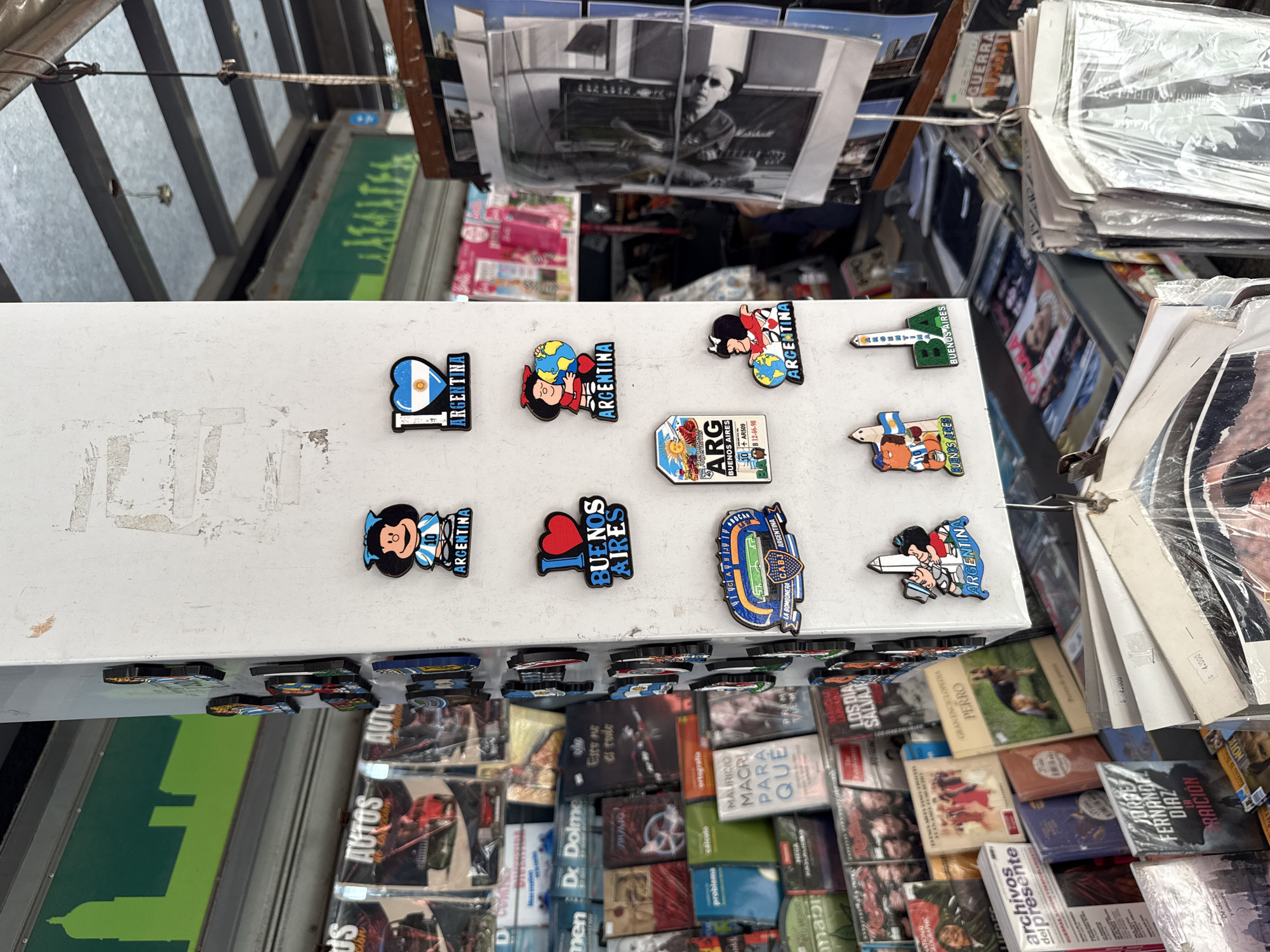 A street vendor in Buenos Aires, Argentina, displays tourist magnets and books for sale. The magnets show the Argentine flag, maps of Buenos Aires, and cartoon characters. Books and magazines are displayed below the magnets and include titles on history, politics, and culture. Photographs are hung above the display.