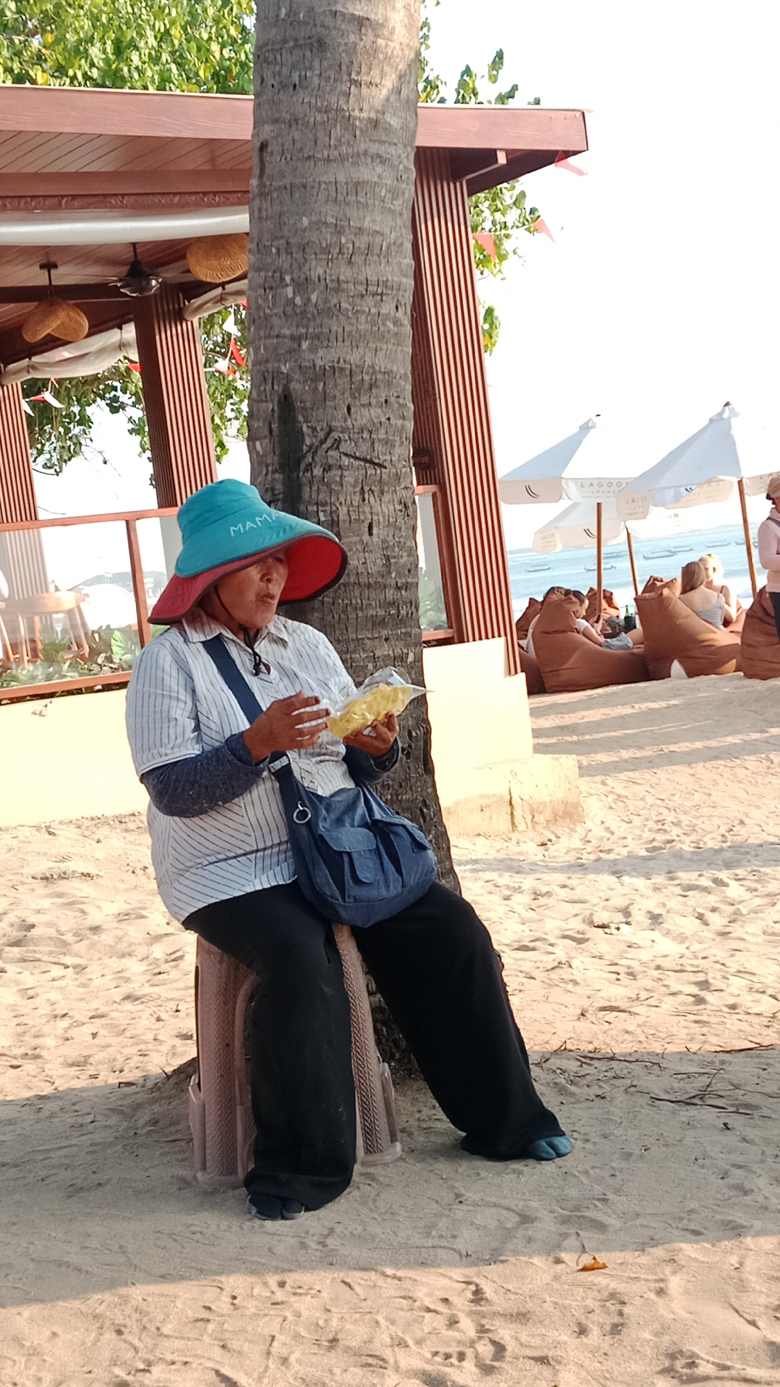 A woman wearing a wide-brimmed hat and a striped shirt sits on a small stool on a sandy beach in Kuta, Indonesia. She is holding a packet of yellow food and appears to be eating it. Her hat is teal with a red brim and the word "MAMA" is visible on the front. She has a blue bag slung over her shoulder. In the background, there are beach umbrellas, beanbag chairs, and other people relaxing. The scene is set near a palm tree and part of a wooden structure, possibly a beachside cafe or bar. The lighting suggests it is late afternoon.