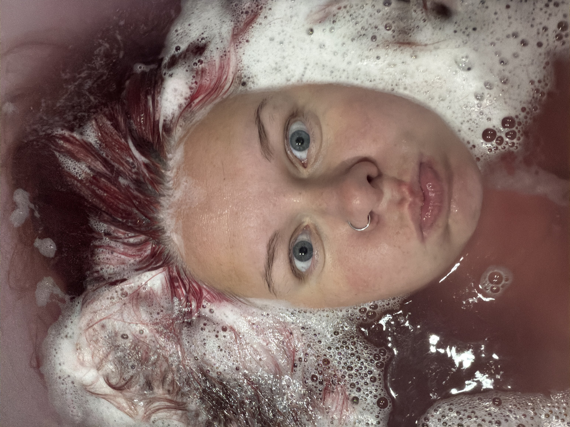 The image is a close-up, overhead shot of a person's face submerged in reddish-brown water and covered in white foam. The person has bright blue eyes and is wearing a silver hoop nose ring. Their mouth is puckered, and strands of reddish-pink hair are visible, mixed with the foam. The water appears to be in a bathtub, indicated by the edge visible in the upper portion of the frame. The overall impression is that of a person taking a bath, possibly with a bath bomb or colored bath product. The location in Chaikovsky, Russia, is not discernible from the image itself.