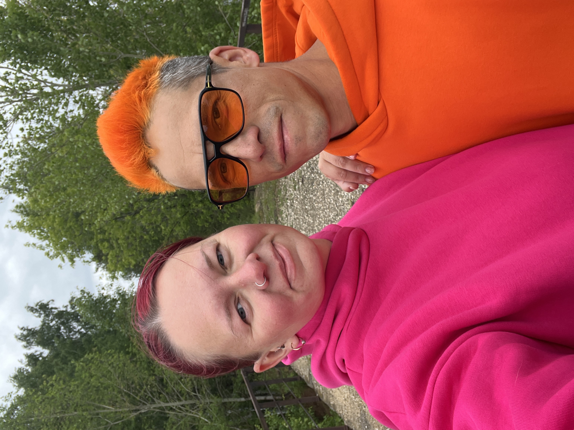 Two people, likely a couple, pose for a selfie outdoors, with trees and an overcast sky visible in the background. The person in the top portion of the image wears an orange hoodie and orange-tinted sunglasses. Their hair is dyed orange at the front, transitioning to gray at the back. The person in the bottom portion of the image wears a pink hoodie and has short, dark pink hair. They have a nose ring and earrings. The image orientation suggests that the camera was held above and tilted. The exact location within the city of Chaikovsky, Russia, cannot be determined from the image alone, as there are no identifiable landmarks.