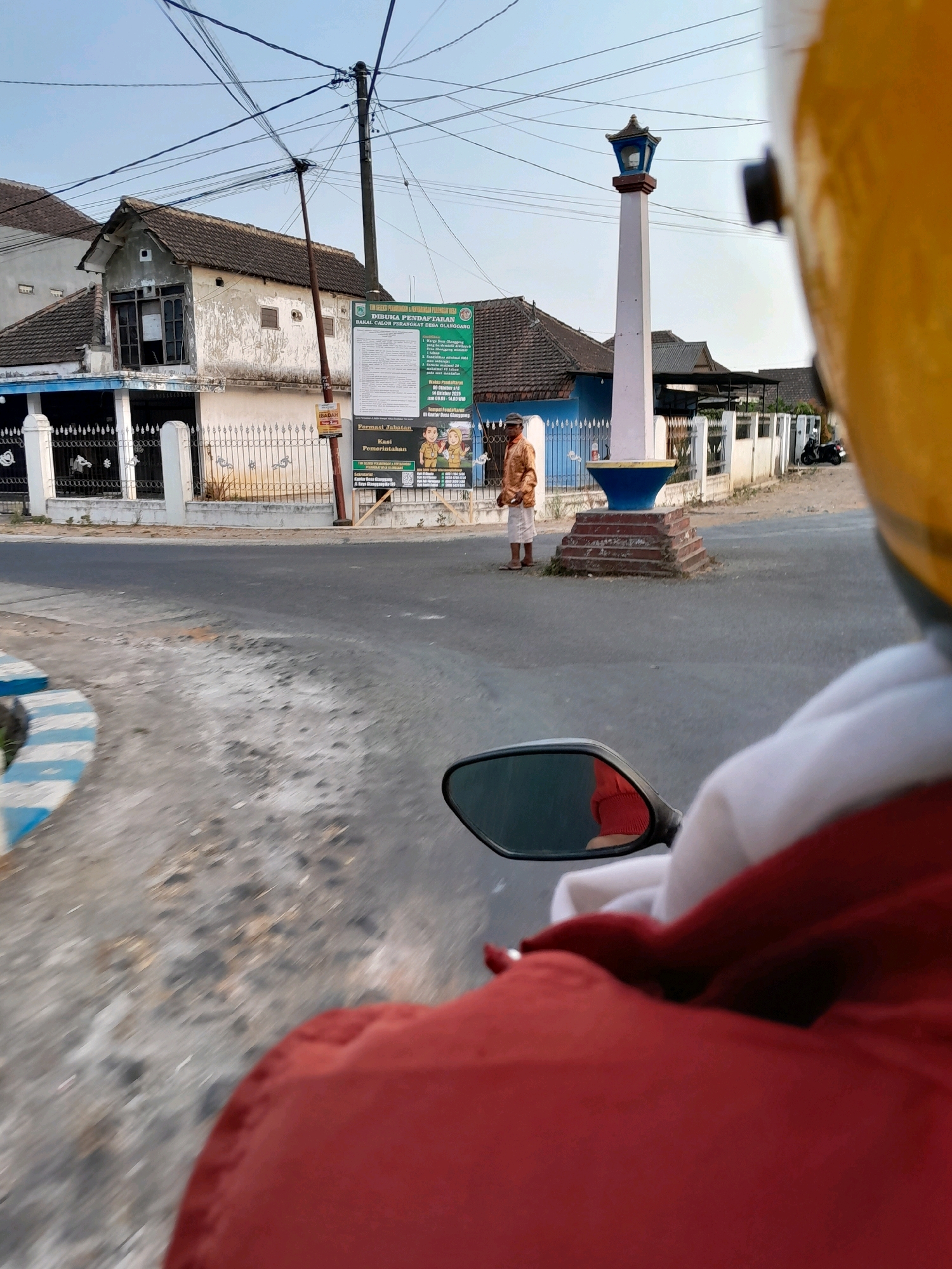 In the city of Jatirejoyoso, Indonesia, a person rides a motorcycle, their yellow helmet and red jacket visible in the foreground. The motorcycle's side mirror reflects the rider's leg and the surrounding asphalt road. In the middle ground, a lone man stands near a monument that features a white obelisk topped with a blue and black lantern. The monument is situated on a raised platform with red brick steps. Behind the monument, a government notice board displays information in Indonesian. The scene is framed by buildings, including a weathered house with an overgrown roof and a row of more modern structures with white fences. Electrical wires crisscross the sky, a common sight in urban and semi-urban areas. The overall environment suggests a public space, possibly a town square or intersection, in an Indonesian city.