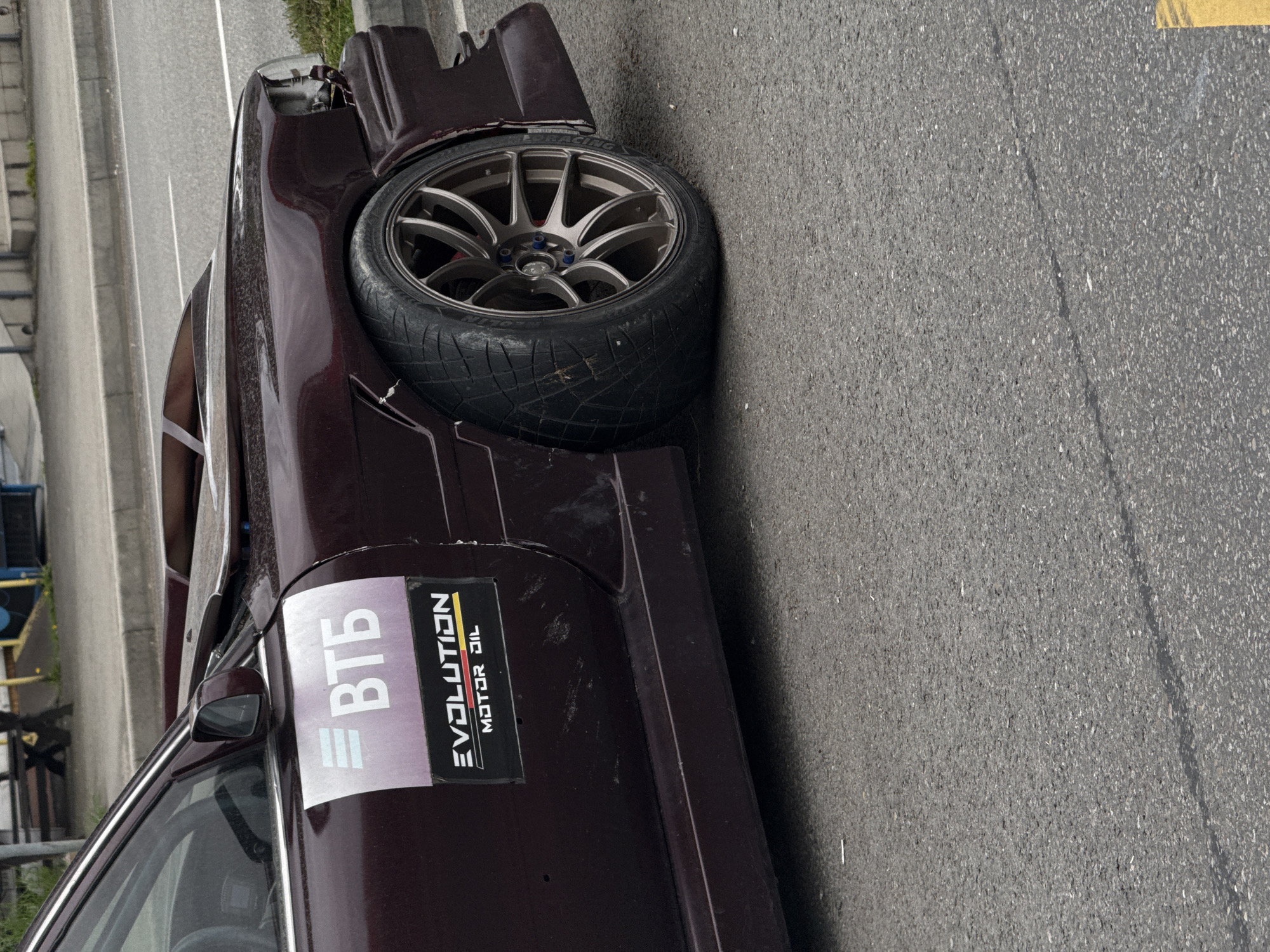 A dark-colored, heavily modified car, possibly a drift or race car, is parked outdoors on asphalt pavement. The image is taken from a side view, looking along the vehicle's length, with the car's front facing towards the top of the frame.
The car's front end appears damaged or in a state of modification, with the bumper removed or severely altered, revealing internal components. The front right wheel and tire are prominently featured. The wheel is a multi-spoke alloy design with a bronze or gunmetal finish, and blue lug nuts are visible. The tire is severely worn, showing extensive performance use with visible cord on its outer edge, and "RACING" text is faintly visible on its sidewall.
On the side of the car, near the door, there are two stickers. One is white with blue/purple text, clearly displaying "ВТБ" (VTB, a Russian bank). The other is a black rectangular sticker with yellow and white text that reads "EVOLUTION MOTOR OIL". The car's bodywork shows some dirt and minor scratches.
The setting is an outdoor area, likely a roadside or parking lot, with grey asphalt visible. In the background, there are concrete barriers or structures and some sparse greenery, suggesting an urban or industrial environment. The lighting indicates it is daytime, possibly under overcast conditions, as shadows are soft. Given the context of Sochi, Russia, and the car's characteristics, it may be associated with a motorsports event or enthusiast gathering.