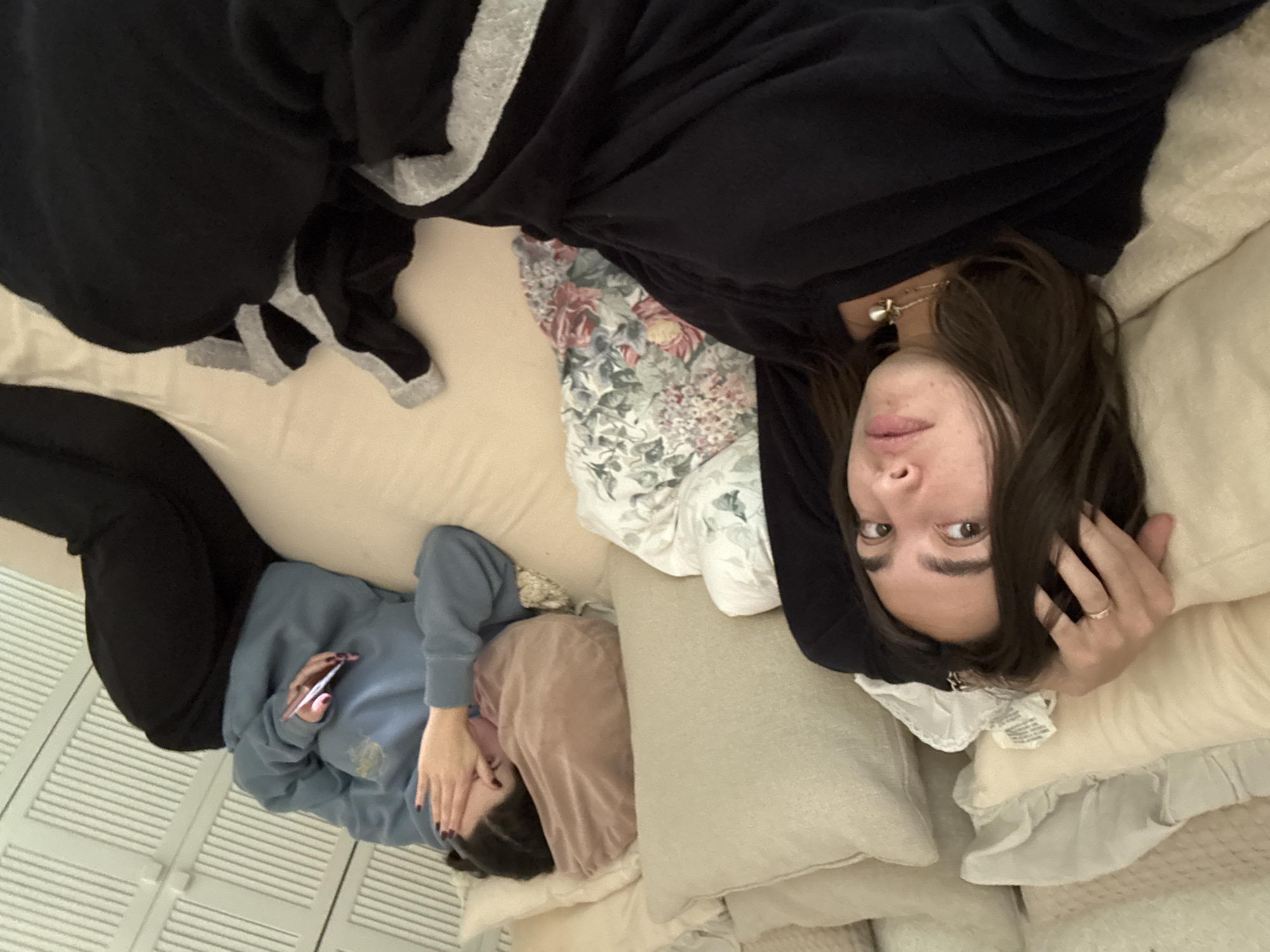 This image displays two individuals casually lounging on a bed or large couch in an indoor setting, likely in Mexico City, Mexico. The scene is captured from an overhead perspective.
In the foreground, a young woman with long dark brown hair lies on her back, resting on light-colored pillows. She is wearing a dark long-sleeved top, possibly with grey trim, a necklace with a small pendant, and a pearl earring. Her eyes are open and directed towards the camera, conveying a neutral or slightly curious expression, with her lips slightly parted. One hand, featuring a ring, is positioned near her head. A small white tag, with unreadable text, is visible near her neck.
Further into the background, another individual is partially visible. They are wearing a light blue long-sleeved sweatshirt and black pants. Their face is largely obscured by their hands, one of which is holding a smartphone with a pink case. Dark nail polish is visible on their fingers, and a light brown or pinkish fabric covers part of their head and shoulder area.
The bedding consists of light-colored sheets and pillows, with a floral patterned sheet or blanket visible between the two people. In the background to the left, a white slatted structure, resembling closet doors or window blinds, can be seen. The overall atmosphere suggests a relaxed and informal moment of leisure or downtime.