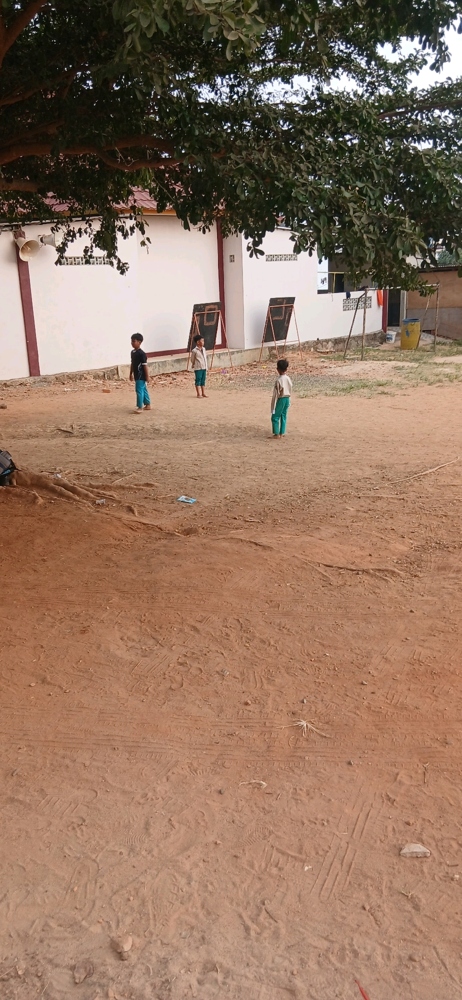 The image depicts an outdoor scene with three young boys in a dirt yard. The boys are standing in front of a white wall with red trim, and two chalkboards on stands are visible. One boy, wearing a black shirt and blue pants, stands to the left. In the center, a boy in a light-colored shirt and blue pants stands near a chalkboard. To the right, another boy in a similar light-colored shirt and blue pants faces away from the camera. Overhead, the branches of a large tree with green leaves create a canopy. The ground is covered in dirt with some scattered debris. A trash can and a swing set are partially visible in the background. The scene appears to be in a schoolyard or a similar outdoor educational space.