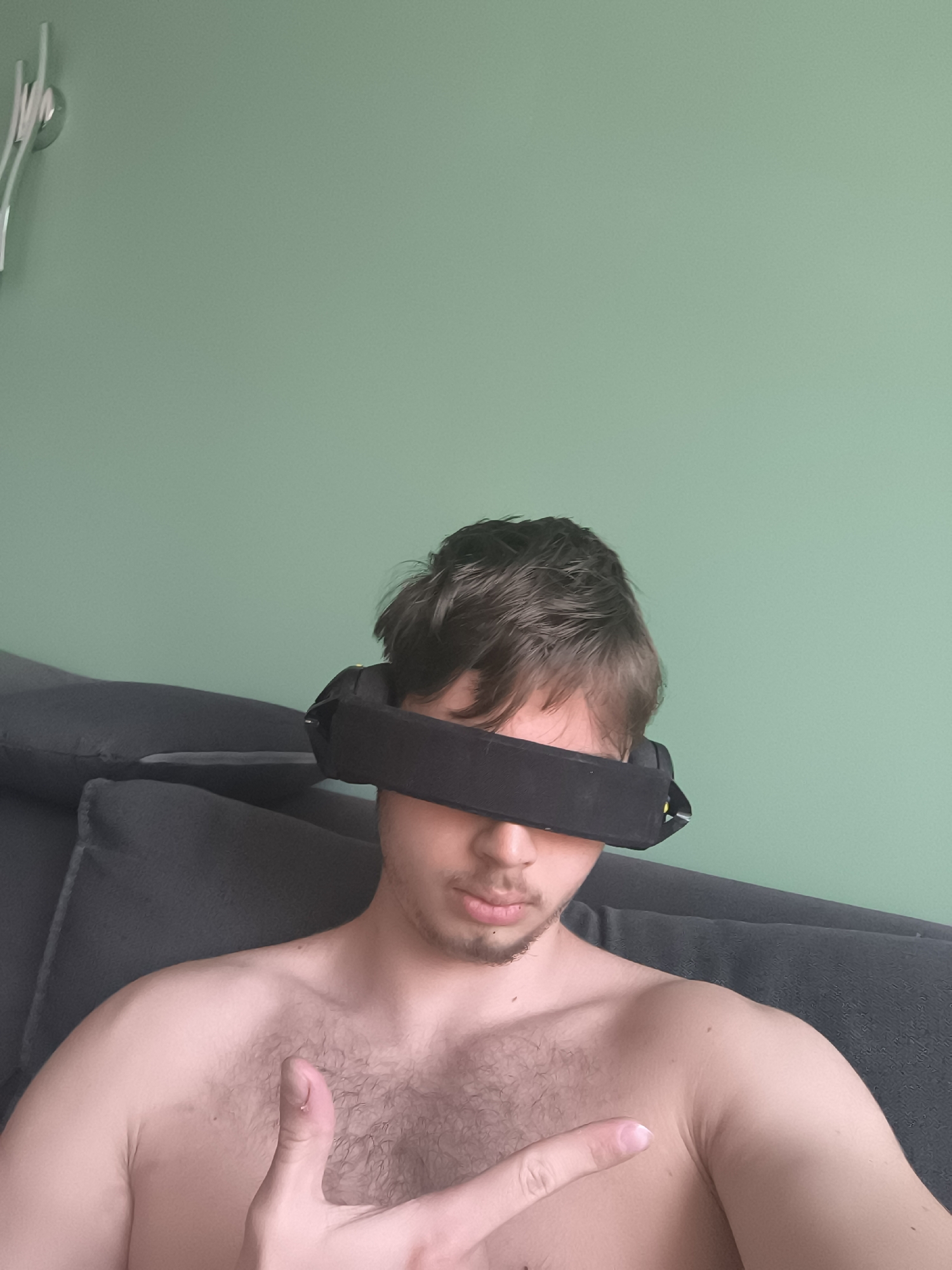 A young man is depicted from the chest up, seated indoors on a dark gray sofa or armchair. He is shirtless, revealing his chest and shoulders, which have some body hair. A wide, black blindfold or sleep mask covers his eyes and forehead, obscuring his upper face. His dark hair is messy and swept back slightly. A light goatee is visible on his chin and upper lip.
His right hand is raised, with his index finger extended and his thumb pointing upwards, forming a gesture that might be interpreted as pointing or a specific hand sign. He appears relaxed, with his mouth slightly ajar.
The background features a plain, light green wall, illuminated evenly, suggesting daytime. In the upper left corner of the image, a portion of a white, abstract decorative object, possibly a clock or wall art, is visible against the green wall. There are no explicit visual cues in the image to indicate the specified location of Sofia, Bulgaria. No text is visible in the image.