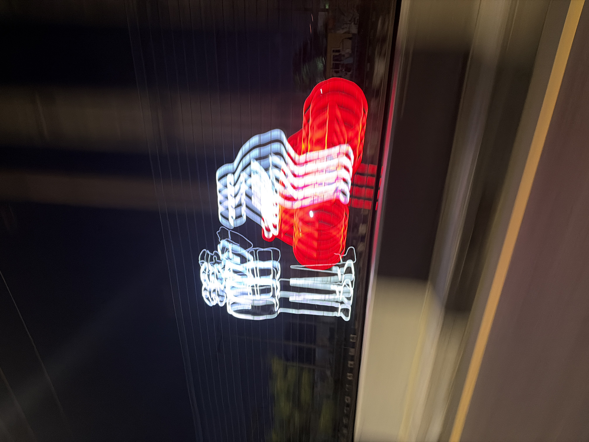 A long-exposure photograph shows a blurred neon sign against a dark background, presumably taken from a moving vehicle. The sign appears to depict a stylized logo or emblem, with red and white elements. The white portion seems to be below the red portion, and both are distorted due to the motion blur. The background is mostly black, with vertical streaks suggesting buildings or structures. To the right, blurred light streaks indicate movement. The sign likely belongs to an establishment in Ikoyi, Nigeria.