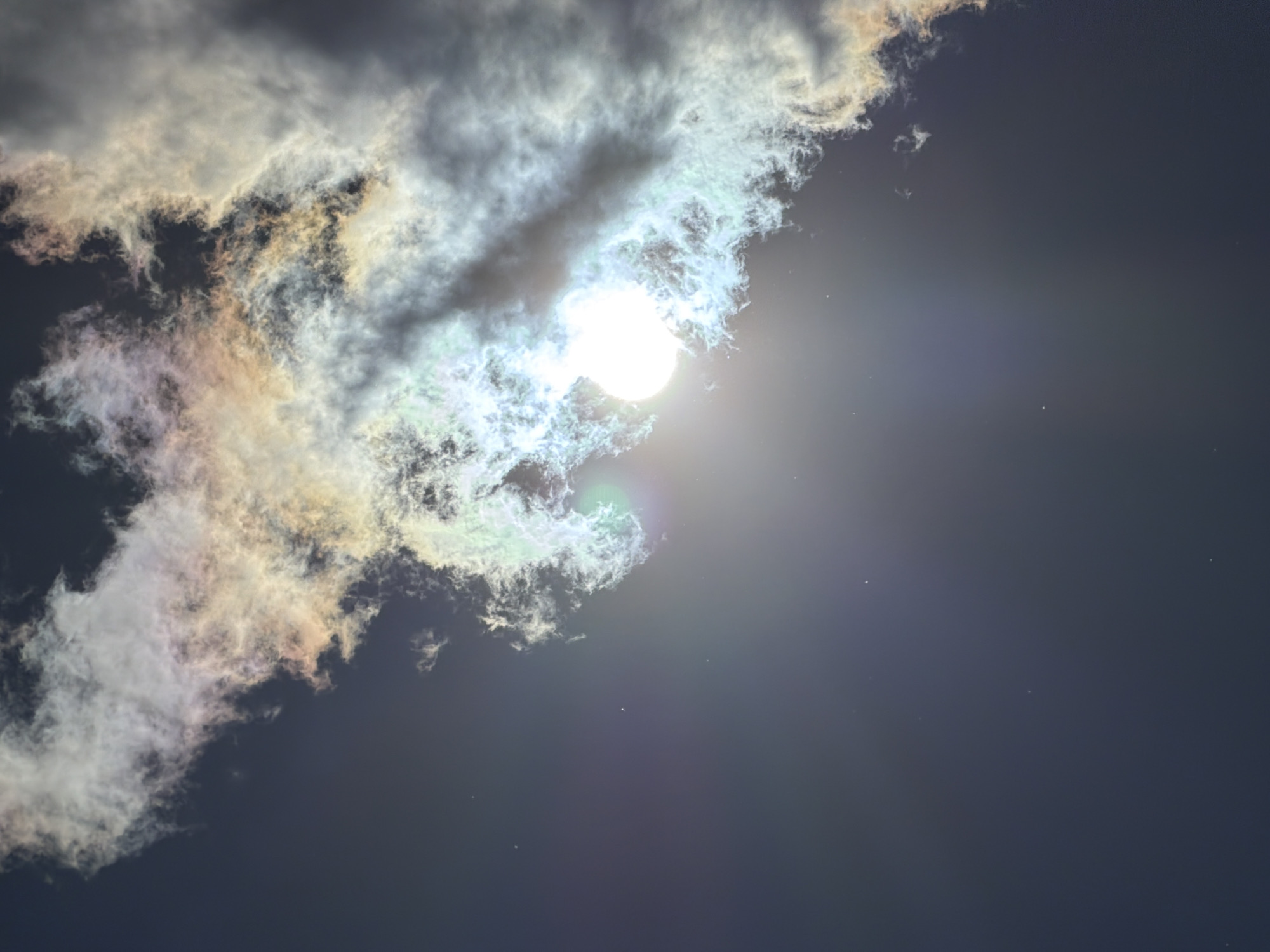 This image depicts a natural phenomenon of iridescent clouds, possibly a form of cloud iridescence or a solar halo, occurring in the sky over Krasnyi Krym, Russia. The sun is partially obscured by these clouds, which exhibit a spectrum of colors, including white, green, yellow, and pink, suggesting the scattering of light by small water droplets or ice crystals within the clouds. The sky itself is a deep blue-gray, indicating it is likely late afternoon or early evening. There are no people or man-made objects visible in the frame, emphasizing the focus on the atmospheric display. The overall environment is one of an open sky, with the clouds occupying the upper portion of the image and extending downwards.