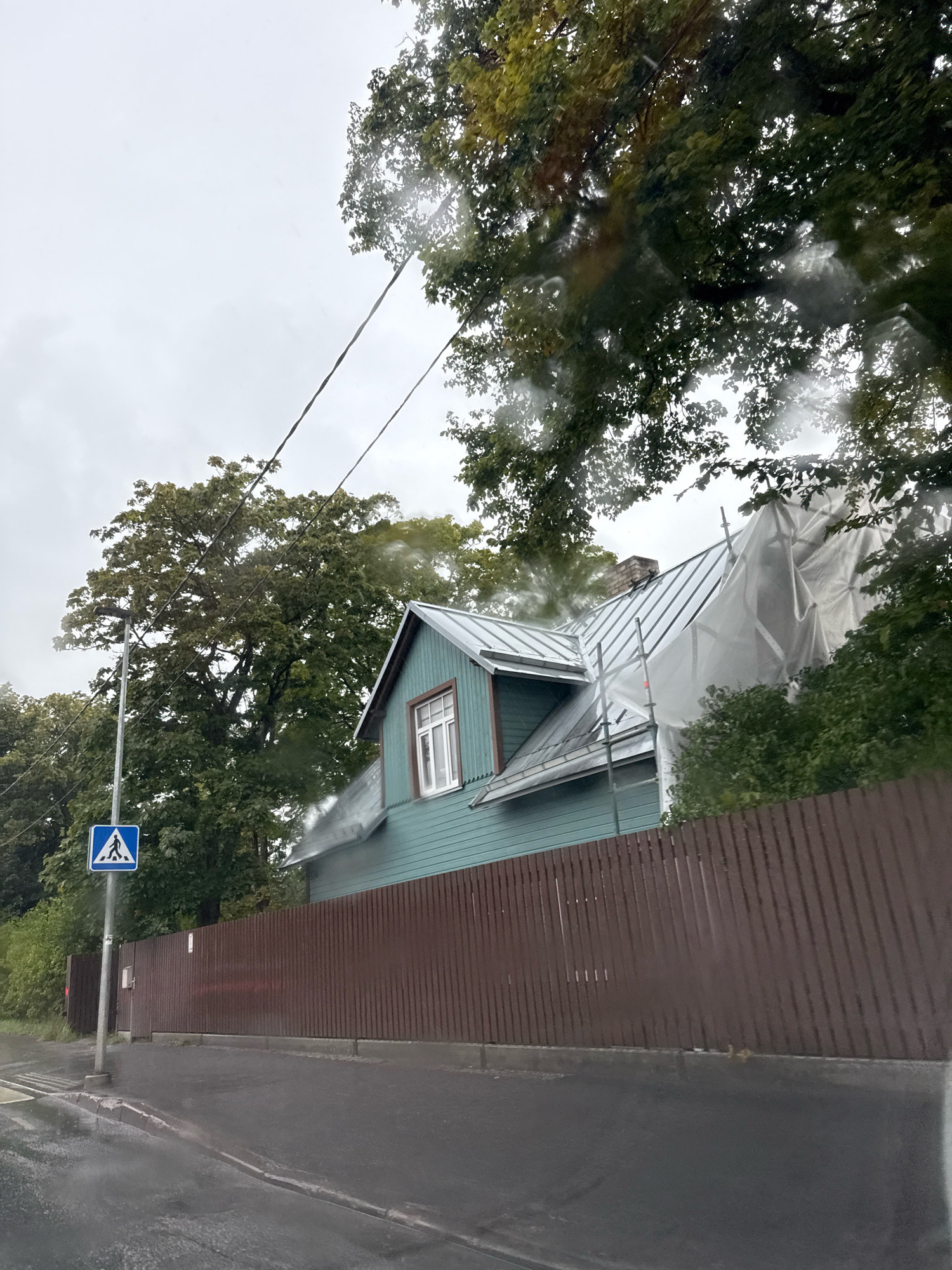 A medium shot captures a partially renovated house on a cloudy day in Tallinn, Estonia. The house, painted teal with white trim, features a light-colored metal roof. Scaffolding surrounds a section of the roof, which is covered with a white tarp. A brown wooden fence runs along the property line, separating it from a wet asphalt road. A pedestrian crossing sign, a blue square with a white pedestrian silhouette, stands on a metal pole to the left of the fence. Trees with green foliage are visible behind the fence and house. The sky is overcast, and there are streaks on the camera lens, suggesting it might be raining or the photo was taken through a wet window.