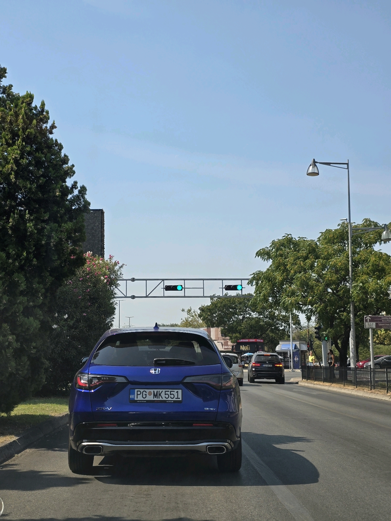 This is a rear-view, eye-level, outdoor, street-level photograph taken on a sunny day in Podgorica, Montenegro. The image captures a traffic scene with a blue Honda ZR-V in the foreground, displaying a license plate that reads "PG MK 551". The car is in motion, moving away from the camera.
Behind the Honda, a black BMW is visible, also in motion. Further down the road, other vehicles and pedestrians are present. Traffic lights are visible, with two of them showing a green signal. The road appears to be a multi-lane asphalt street with painted lane markers.
On the left side of the street, lush green trees and flowering bushes line the sidewalk. On the right side, there are more trees, some buildings with businesses, streetlights, and a black metal fence. The sky is clear and blue. The general impression is a typical urban street scene in Podgorica during daylight hours.