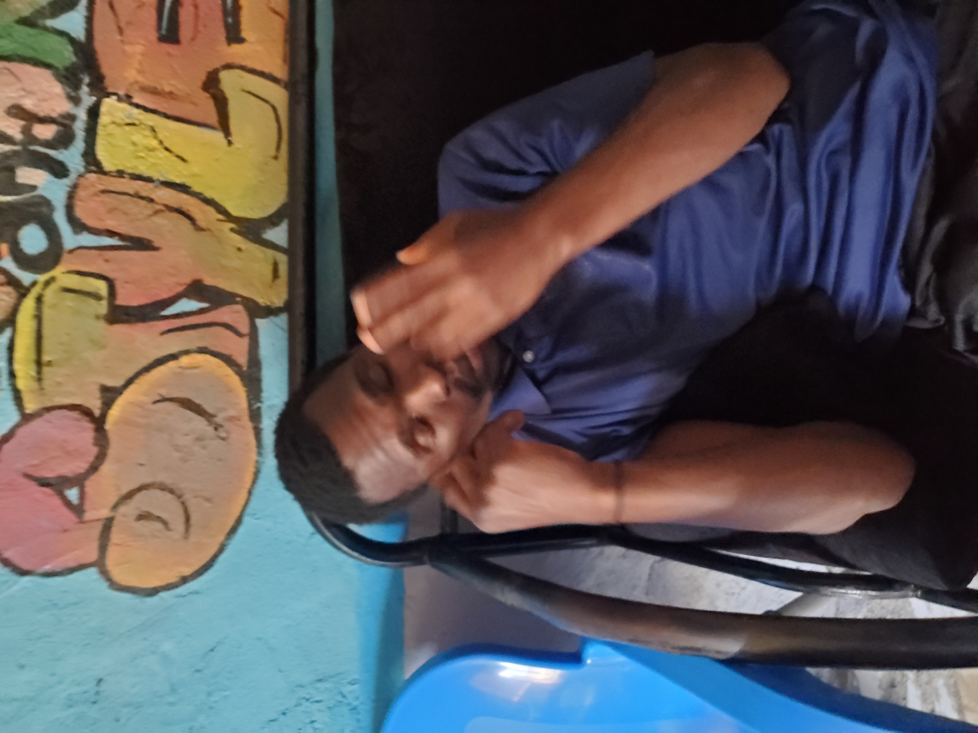 A Black man is sitting on a dark chair with his eyes closed, leaning back, and his head tilted to his right. He is wearing a dark blue polo shirt. His left arm is bent and his hand is touching his ear, while his right arm is bent and his hand is raised near his face. In the background, there is graffiti on a light blue wall with colorful lettering that appears to spell out "STYLE." To the left of the man, a bright blue plastic chair is visible. The scene is likely indoors, possibly in a public space or a private residence in Jos, Nigeria.