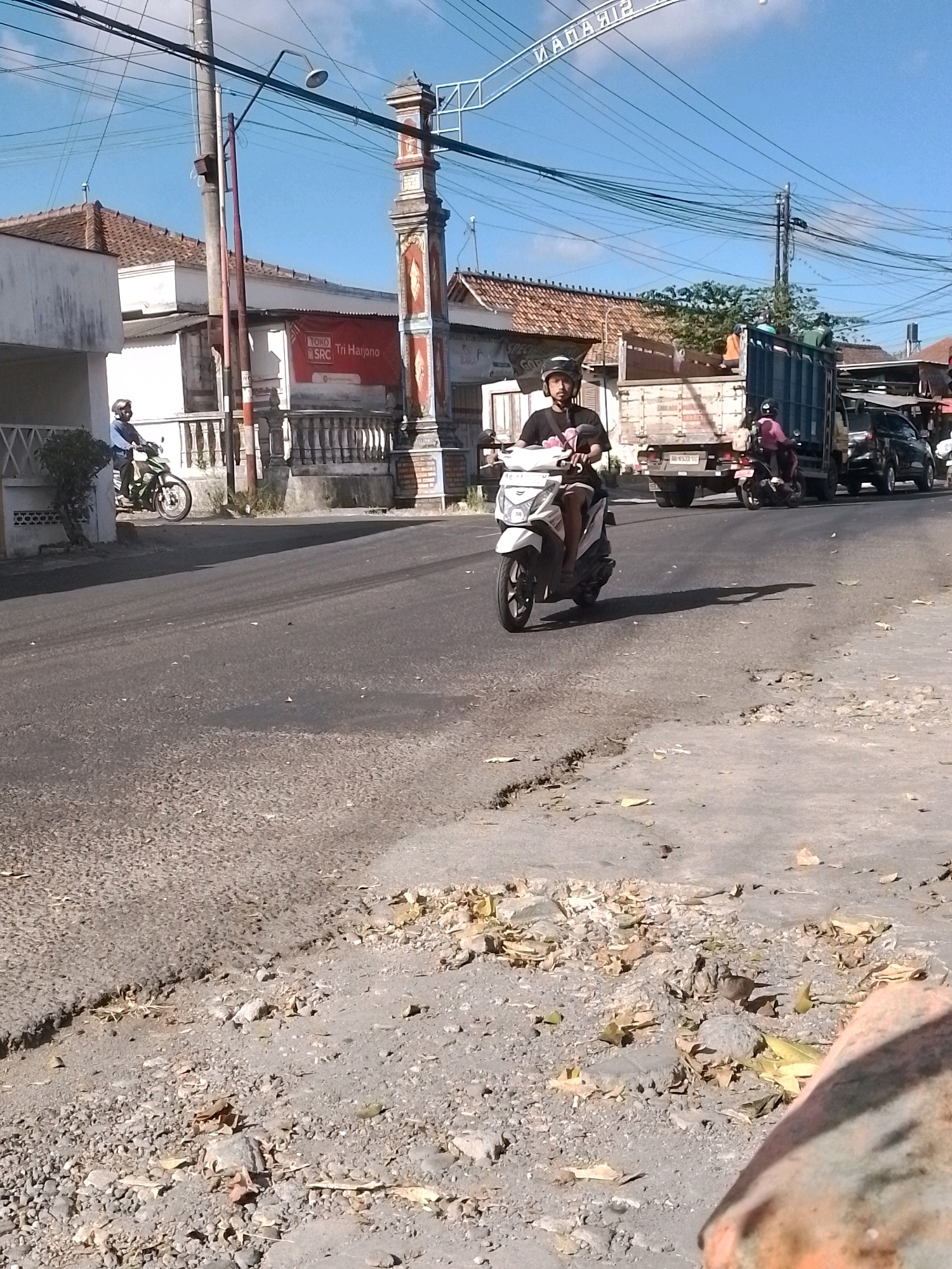 The image depicts a street scene in Wonosari, Indonesia, under a clear blue sky. In the foreground, the asphalt road is uneven with cracks and debris, including fallen leaves and small stones. A white scooter with a rider wearing a black helmet and a dark t-shirt is in the middle of the road, casting a distinct shadow. Further down the road, a large blue truck is parked, and behind it, a black car is visible. Another scooter with a rider in a pink shirt is also present on the road. On the left side of the street, a white building with a tiled roof and a decorative fence is visible. A red banner with text and logos is prominently displayed on the building. A pedestrian walkway with railings is also present. On the right side of the image, a decorative archway or pillar with intricate designs stands near the road. Power lines crisscross the sky above the scene.