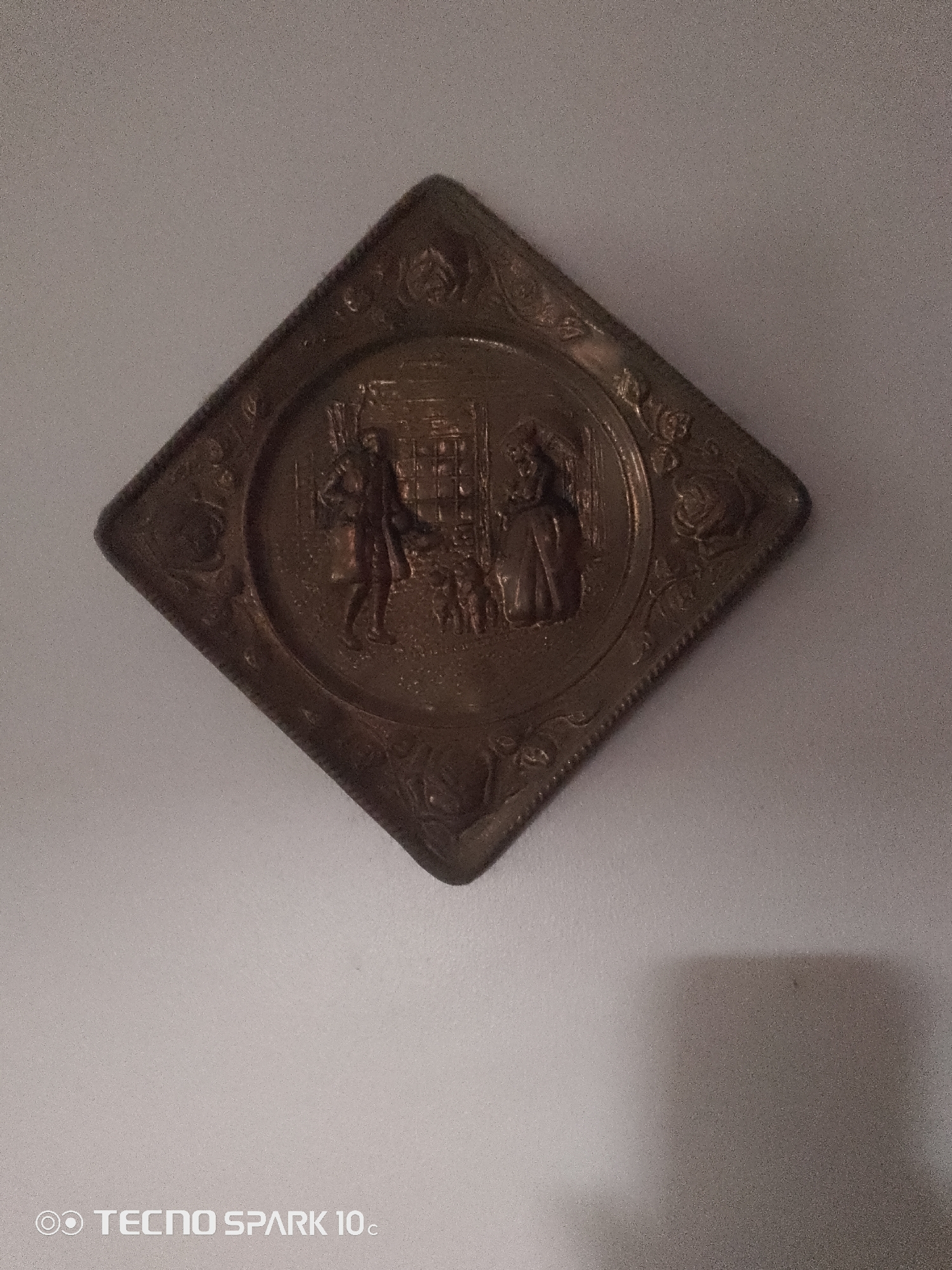 A tarnished, diamond-shaped metallic plate hangs on a pale gray, textured wall, likely in Lekki, Nigeria. The plate's surface depicts a relief scene featuring two figures, a man and a woman, standing near what appears to be a fireplace or hearth. The man, on the left, is depicted in profile, wearing attire that suggests historical clothing. The woman, on the right, is also in profile, dressed in a long, full gown. Between them, a small animal, possibly a dog, is visible. The background within the plate shows a suggestion of interior architecture. The metallic surface of the plate shows signs of age and wear, with a dull patina. The plate is suspended from the wall at a 45-degree angle. The lighting creates a subtle shadow on the wall to the right of the plate. The bottom left corner of the image contains a watermark that reads "TECNO SPARK 10c."