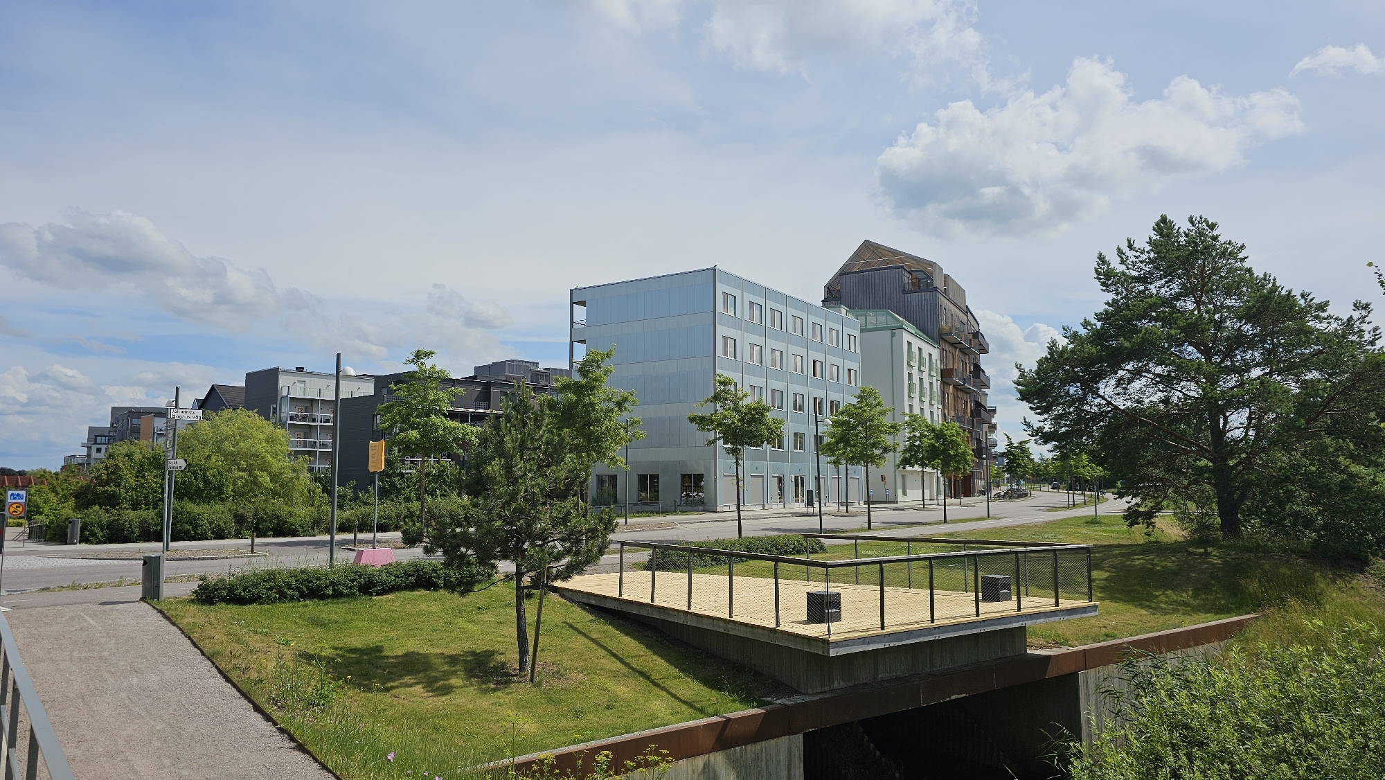 The image captures a scene in Linköping, Sweden, specifically focusing on the area surrounding the Valla Canal, near the Linköping University. In the foreground, a newly constructed wooden deck with a metal railing sits elevated, appearing to overlook a canal or waterway below. Green grass and young trees surround the deck, and beyond that, a concrete path and an access ramp lead away to the left.
Behind the deck, a line of modern apartment buildings stretches across the mid-ground. The buildings are varied in architectural style and color, with some featuring light blue, light green, and darker grey facades. Balconies are visible on some of the structures. A tall evergreen tree stands prominently on the right side of the frame, with its branches extending towards the buildings and the sky.
In the background, more apartment buildings and trees are visible, suggesting a residential or mixed-use urban development. The sky is partly cloudy with white cumulus clouds against a blue backdrop. Street signs are visible, including one with Swedish text "Högskolevägen," indicating the presence of a road named Högskolevägen, which is adjacent to the university and canals in Linköping. A traffic sign with a blue circle and a red border, common in Sweden, is also present. No people are visible in the image. The overall impression is one of a modern, well-maintained urban environment with green spaces.