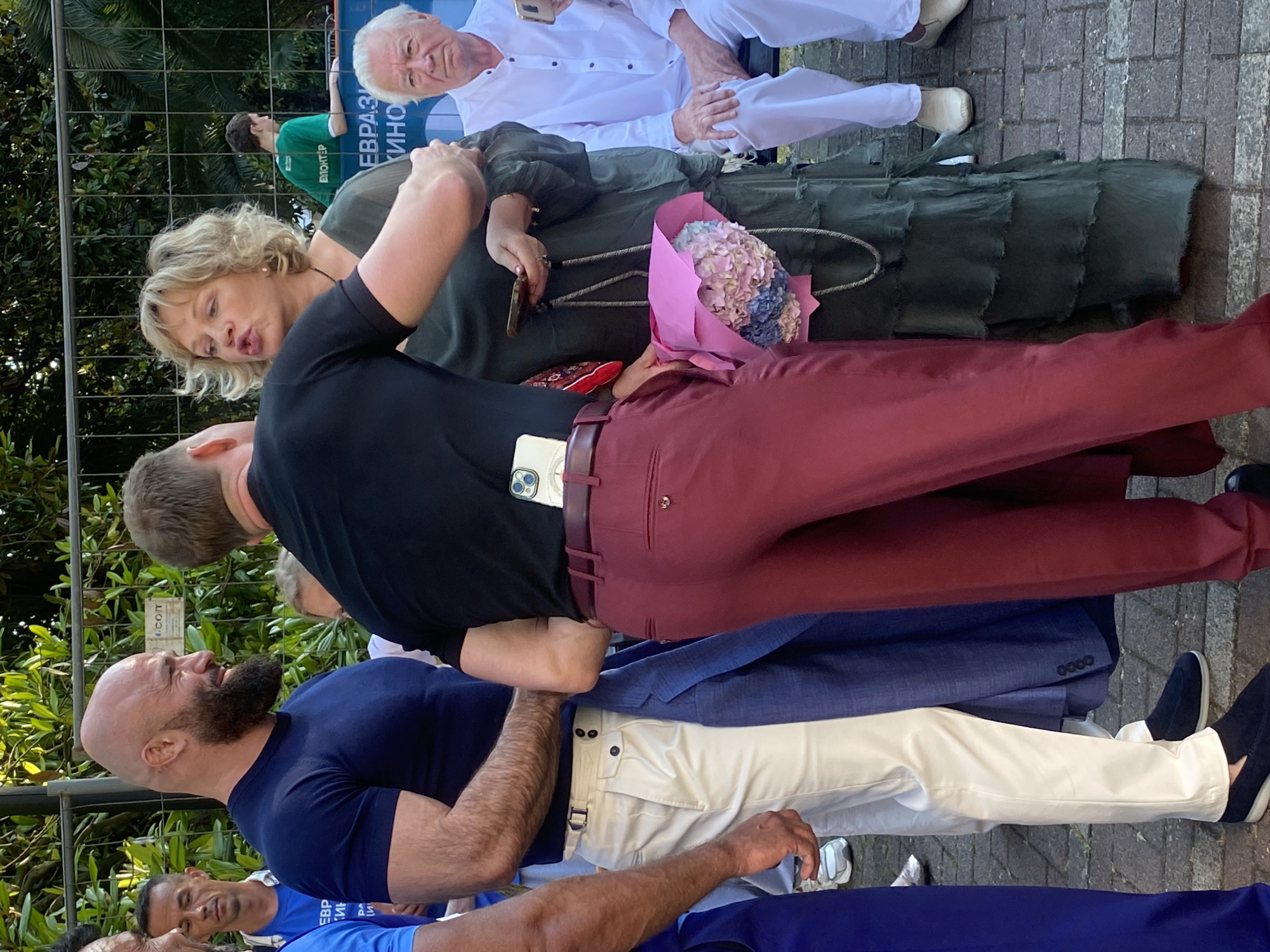The image depicts an outdoor scene in the city of Sochi, Russia. In the foreground, a bald man with a beard, wearing a dark blue t-shirt, stands to the left. To his right, another man, seen from the back, is wearing a black t-shirt and maroon pants. He is embracing a woman with blonde curly hair, who is wearing a dark green dress and holding a bouquet of flowers. Behind them, an elderly man in a white shirt and pants sits on a bench. The background shows a metal fence and lush green foliage. The ground is paved with interlocking grey stones.