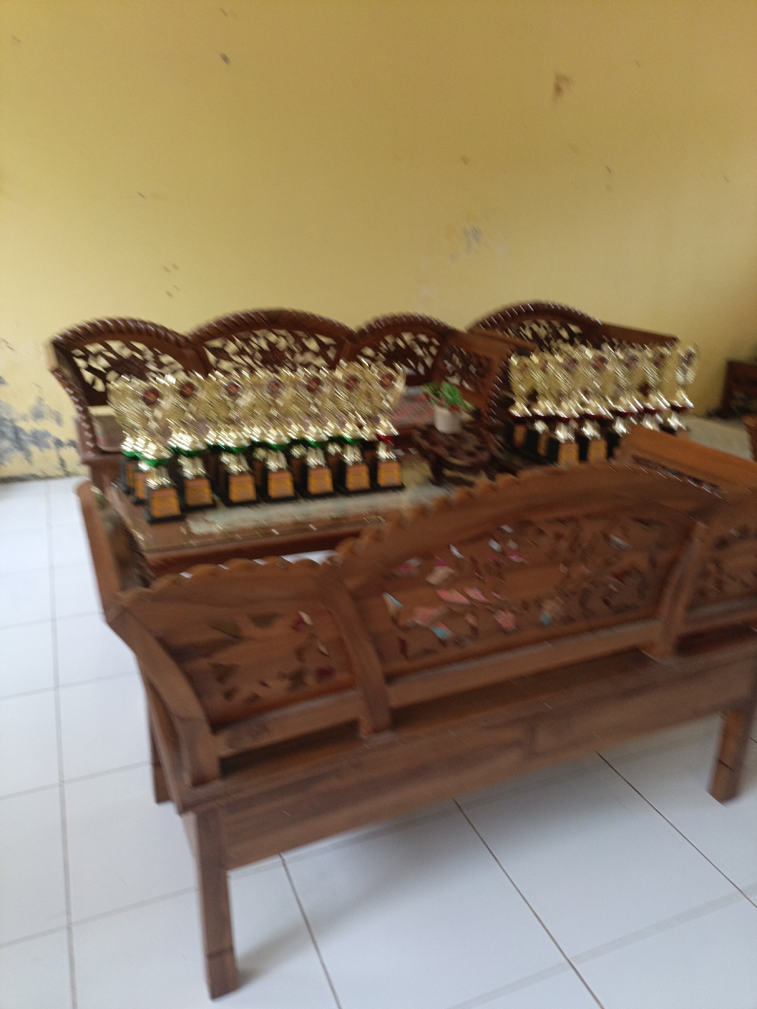 The image shows a wooden bench in front of a set of trophies displayed on another wooden structure. The bench is carved with intricate patterns, and the trophies are gold-colored with red accents. The scene appears to be indoors, with a light-colored tiled floor and a yellow wall in the background. The lighting is somewhat dim, and the image quality is blurry, suggesting it was taken with a low-resolution camera or in low light conditions. The trophies are arranged in rows, suggesting a competition or award ceremony. The presence of the bench and trophies implies a formal setting, possibly a school, community center, or a place of recognition. Without further context, it is difficult to definitively pinpoint the exact location within Lhokseumawe, Indonesia, but it is likely a public or semi-public space dedicated to events or displays.