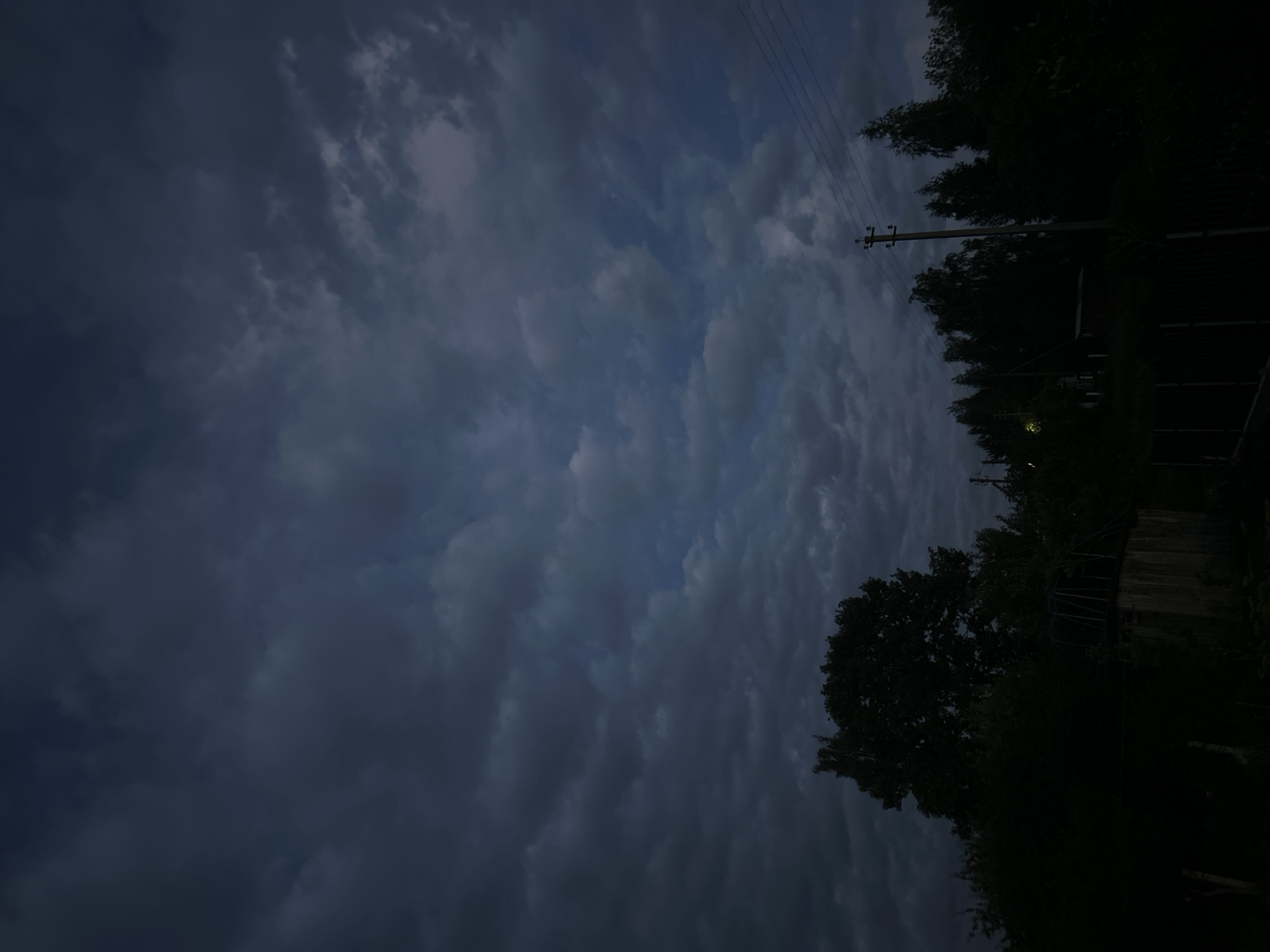 The image depicts a dark, cloudy sky over a rural landscape at dusk or night. In the foreground, silhouetted trees and foliage obscure most of the ground. A small wooden structure, possibly a shed or outbuilding, is visible on the left side. A utility pole with power lines stretches across the right side of the frame, disappearing into the dense vegetation. The overall impression is one of twilight in a secluded, possibly agricultural area within the Забалоцкі сельскі Савет, Belarus.