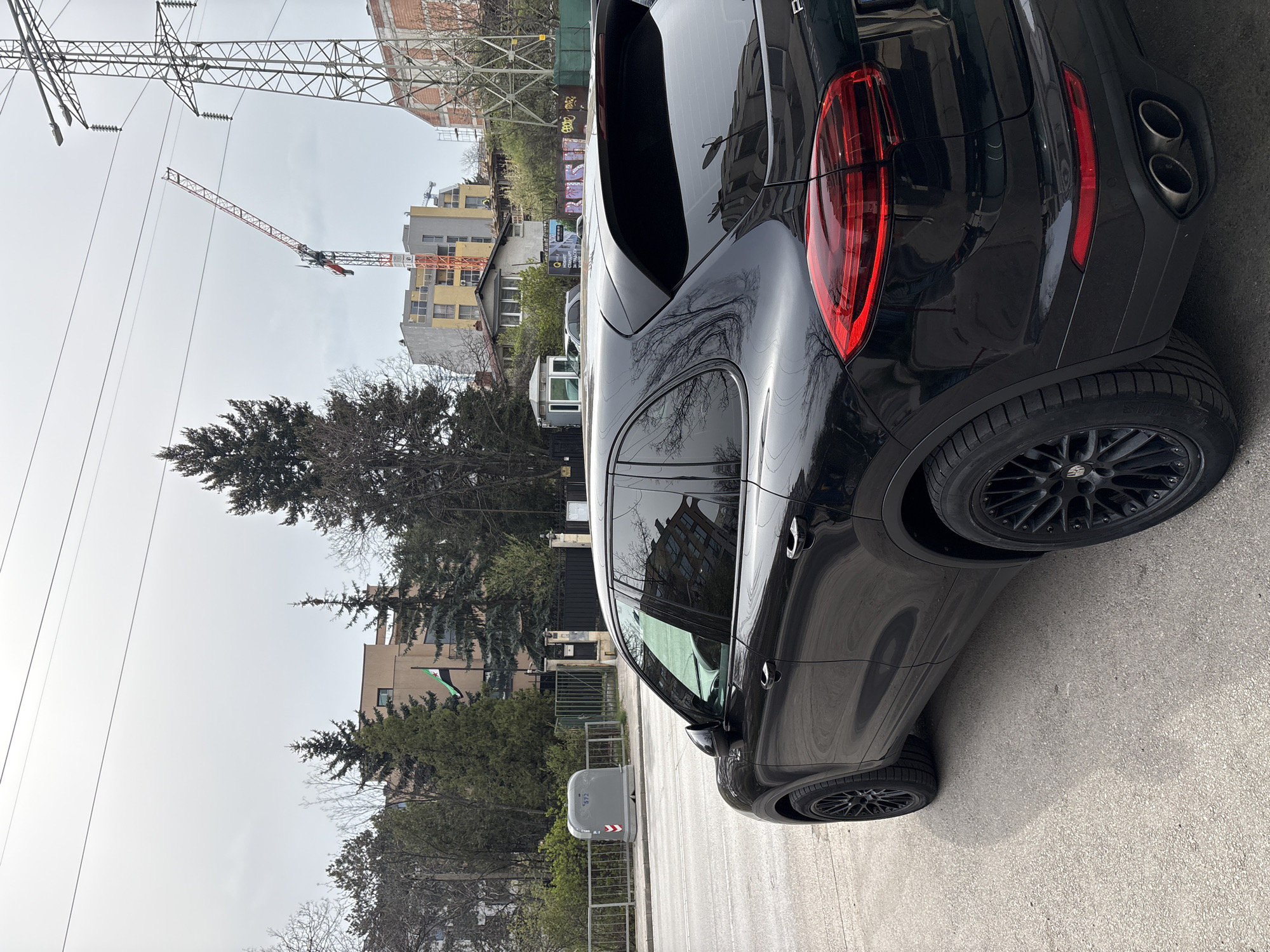 The image depicts a black SUV parked on a street in Sofia, Bulgaria. In the background are residential buildings, trees, power lines, a crane, and a building under construction. A Palestinian flag is visible next to the SUV. There are no people visible. The environment appears to be urban and overcast. A grey utility box stands on the curb near the vehicle.